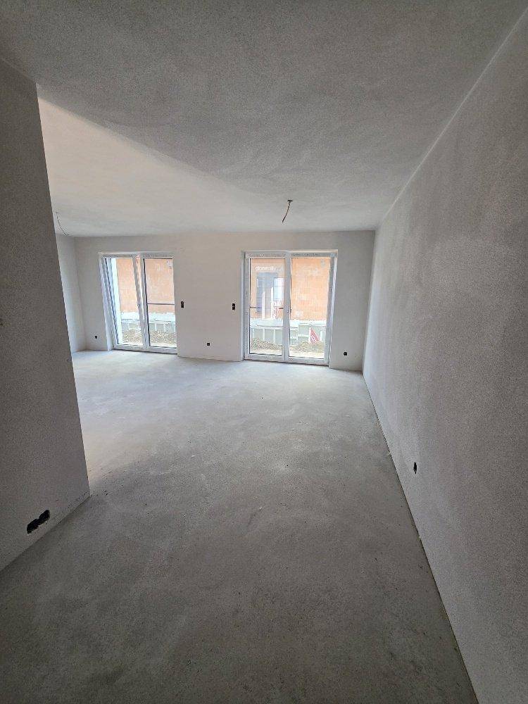 Geräumiger, unfertiger Innenraum mit Betonboden und großen Fenstertüren, die viel Tageslicht hereinlassen.