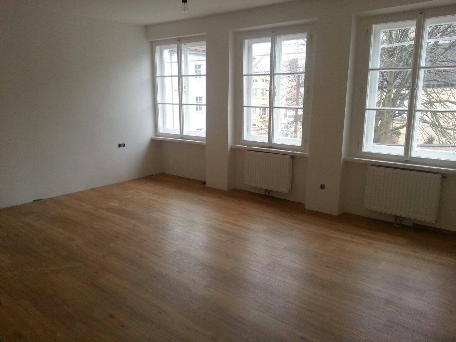 Heller, leerer Raum mit Holzboden und drei großen Fenstern, ideal für Wohnzimmer.