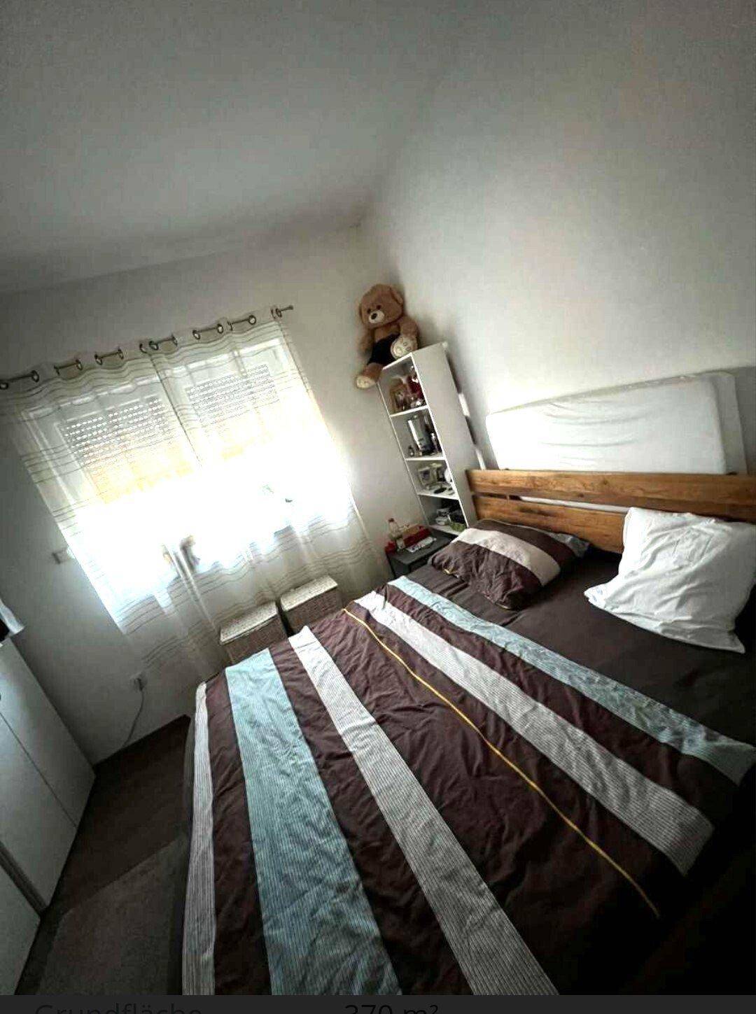 Helles Schlafzimmer mit Holzbett, Nachttisch und Fenster, das viel Tageslicht hereinlässt.