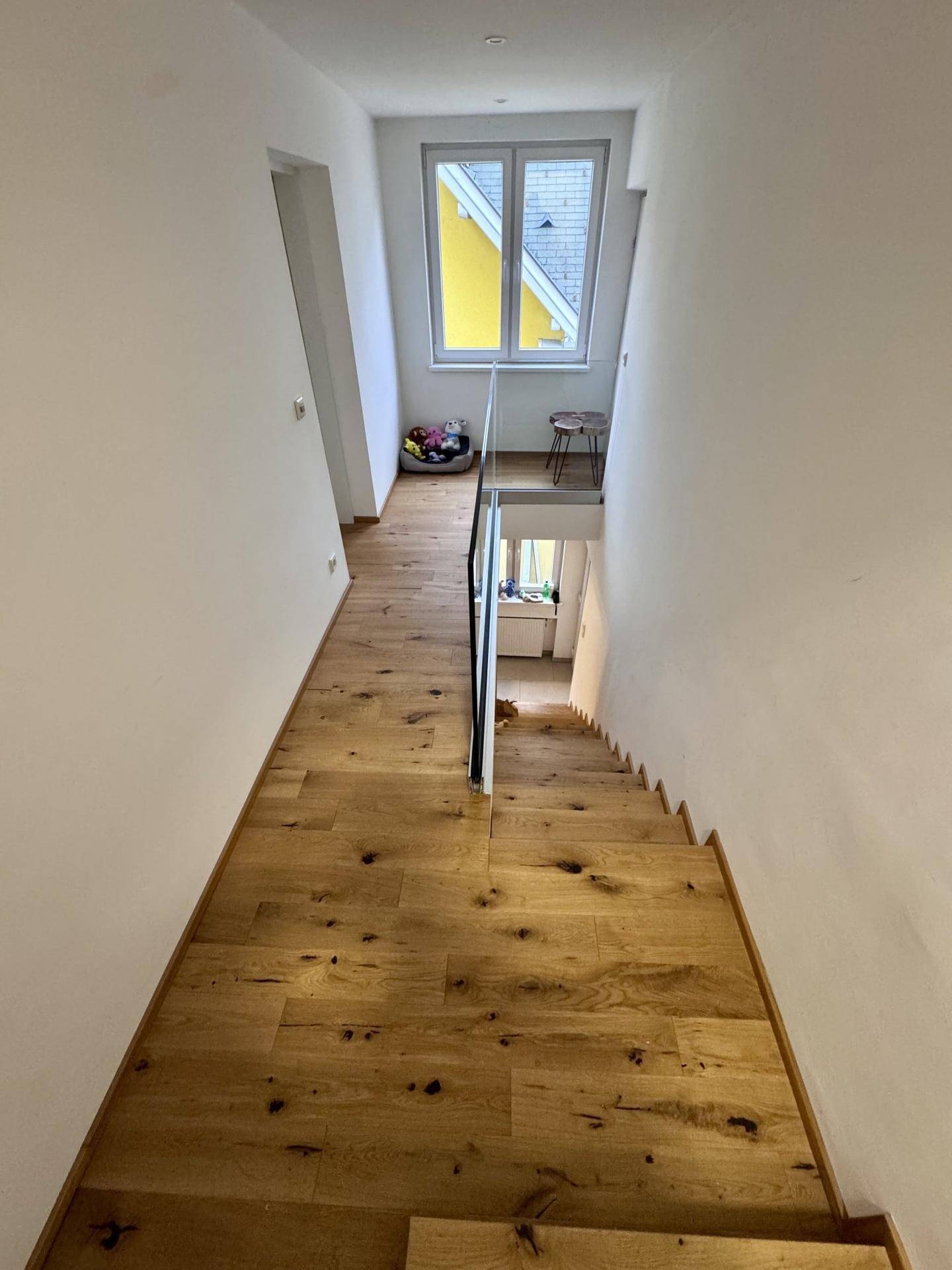 Oberer Treppenbereich mit Holzboden, moderner Glasbrüstung und einem Fenster für natürliches Licht.