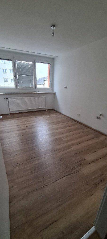 Helles, leeres Zimmer mit Laminatboden, weißen Wänden und großem Fenster.