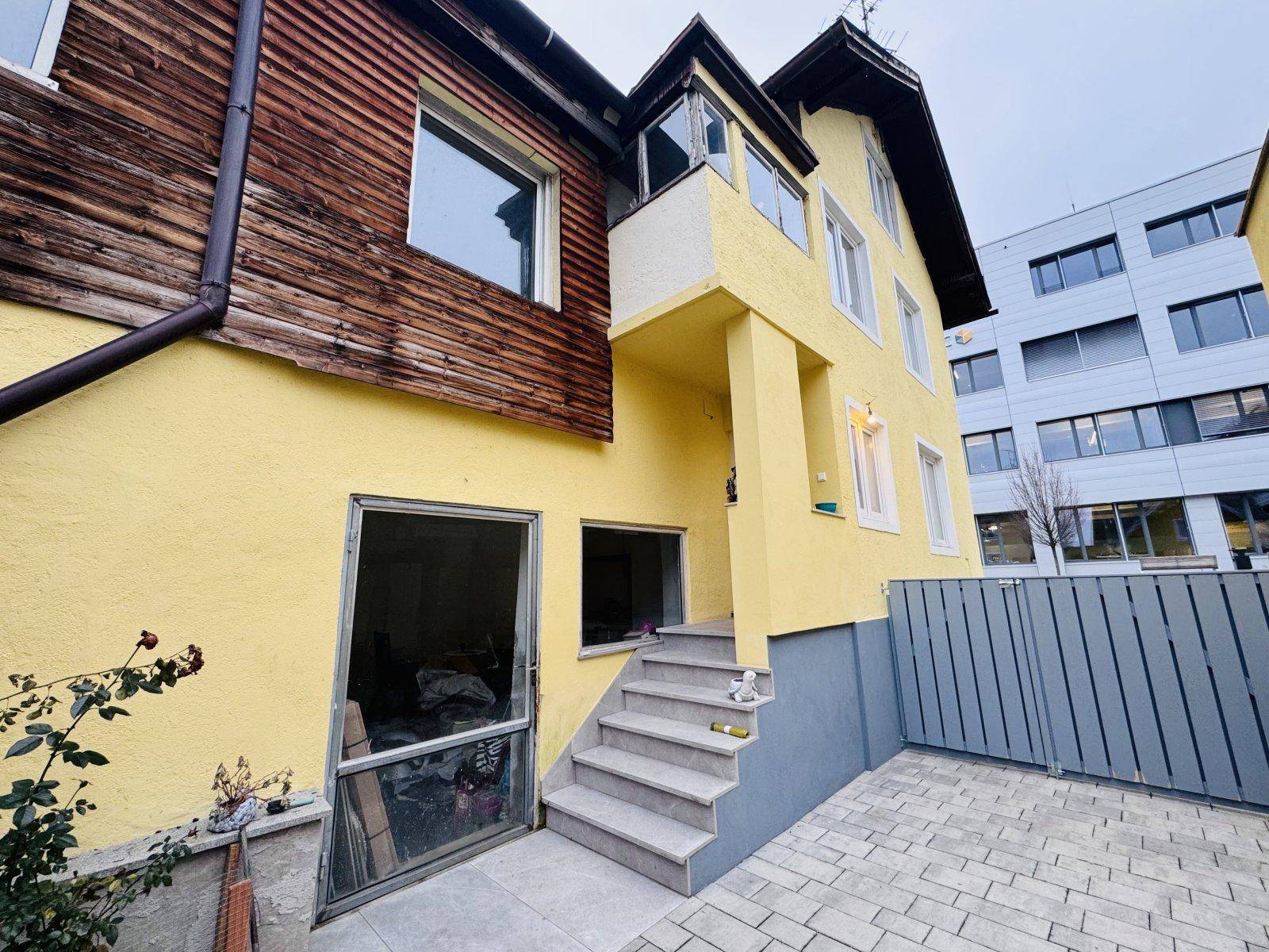 Rückansicht des Hauses mit Außentreppe, Garagenzugang und einem modernen Gebäude im Hintergrund.