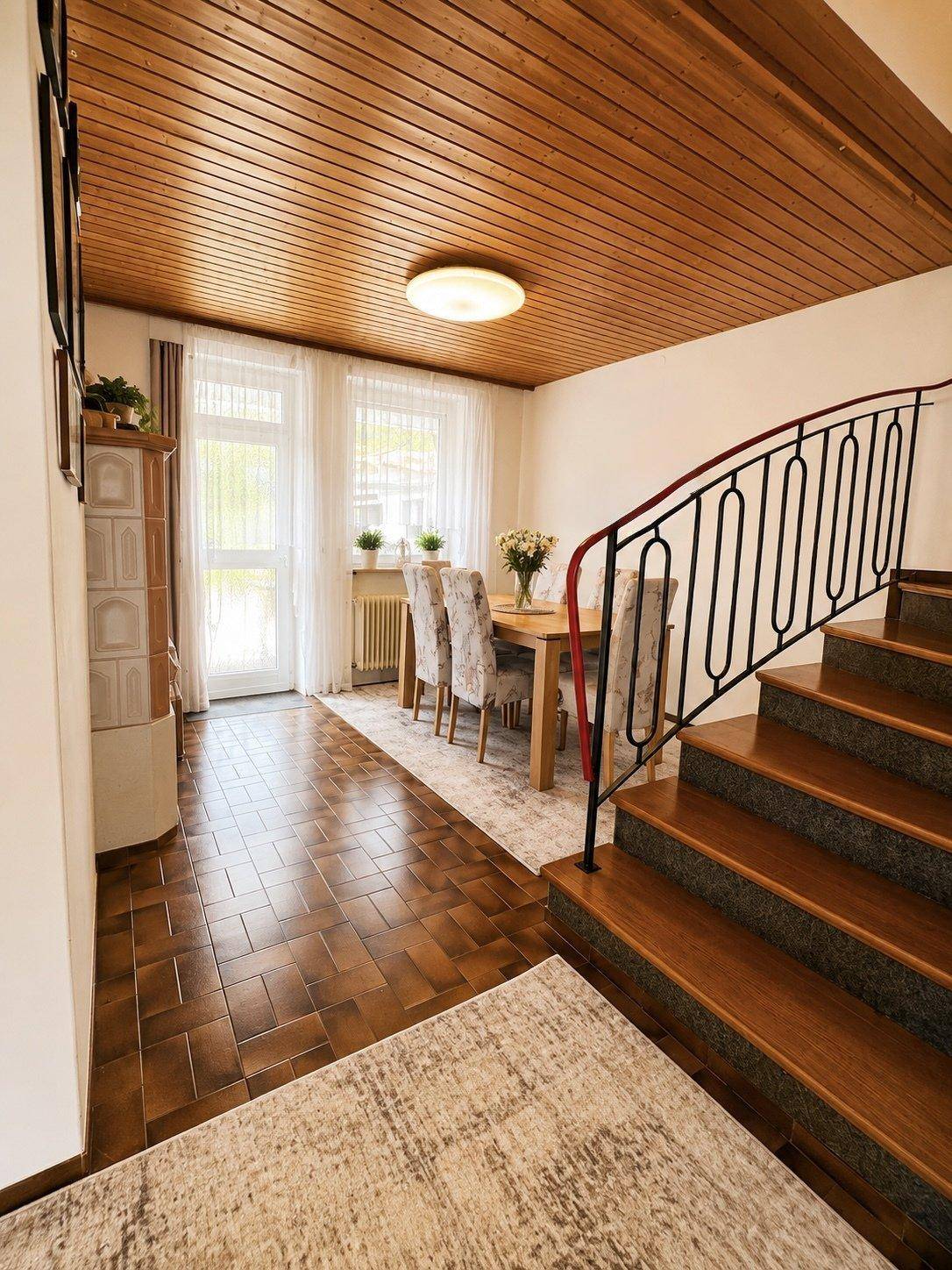 Essbereich mit Holztisch, Kachelofen, Holztreppe und Zugang zum Außenbereich.