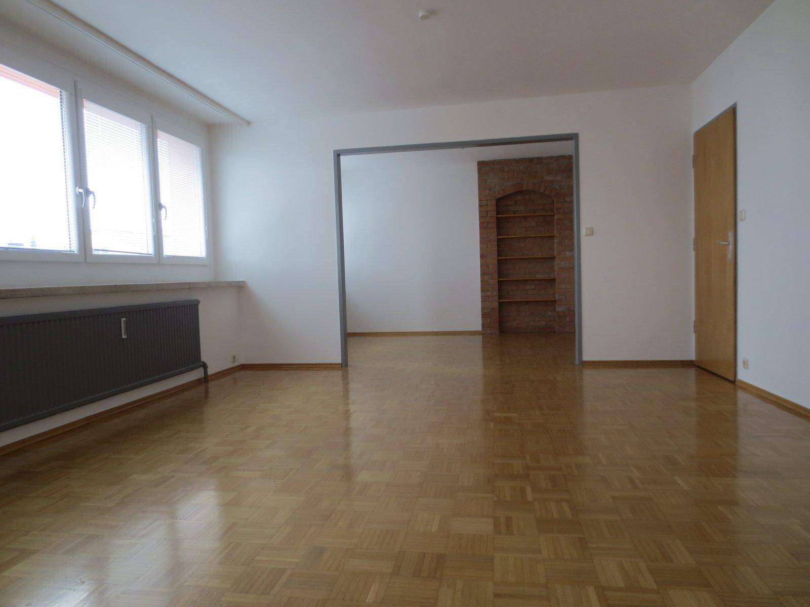 Heller Wohnbereich mit Parkettboden, großen Fenstern und offenem Durchgang zu einem Raum mit Ziegelwand.