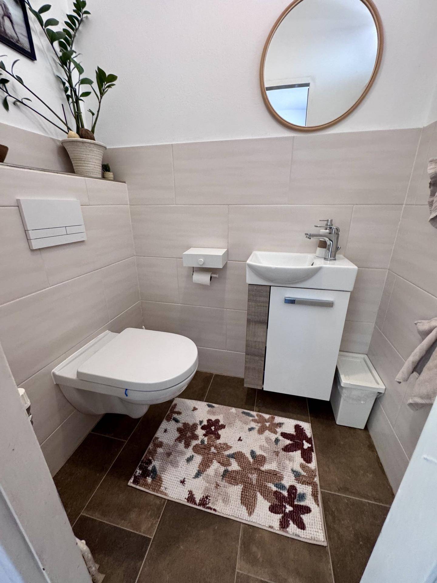 Modernes Gäste-WC mit Wand-WC, kleinem Waschbecken, Spiegel und dunklen Bodenfliesen.