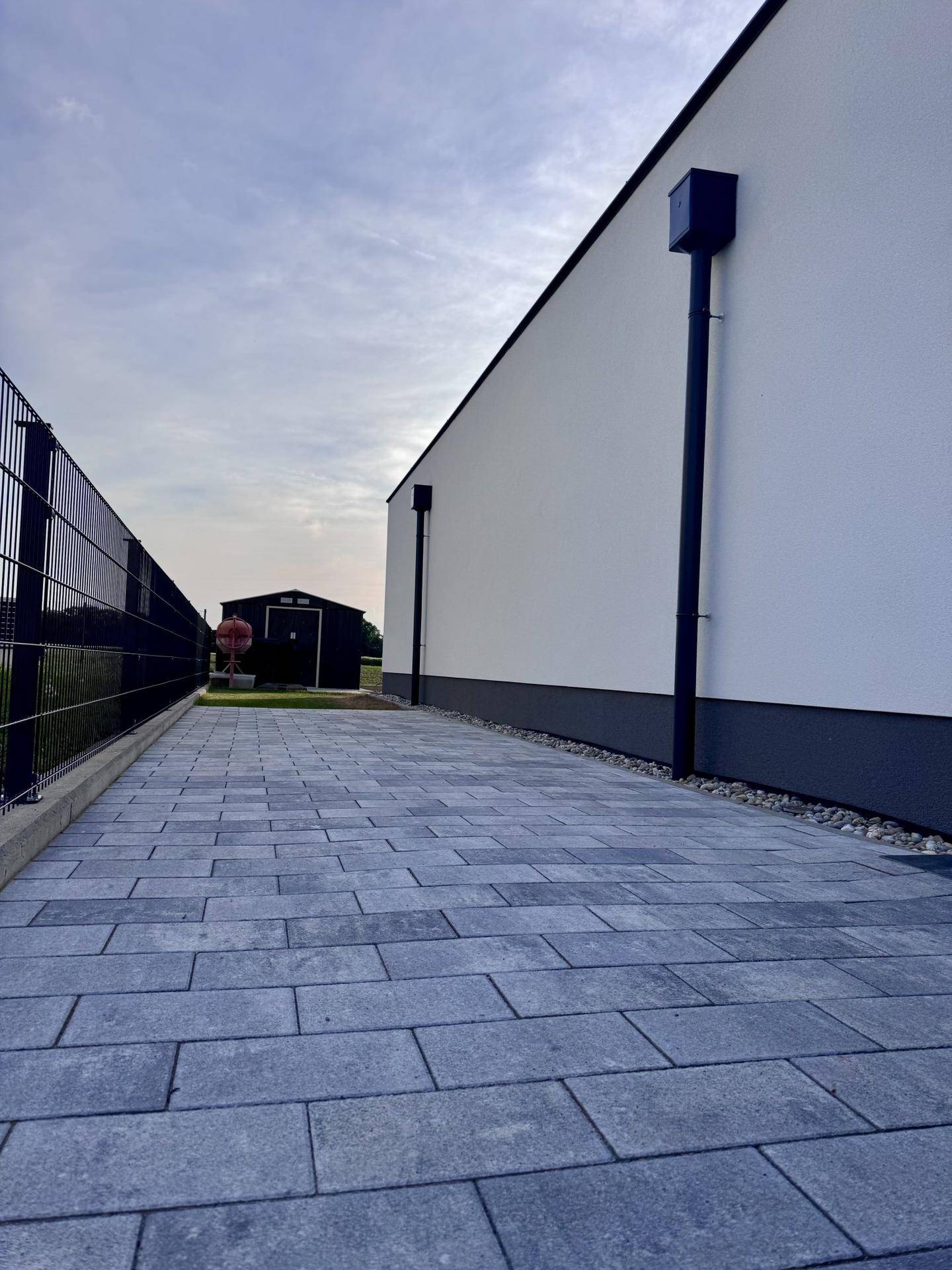 Gepflasterter Weg entlang der Hauswand mit Blick auf ein Nebengebäude im Hintergrund.