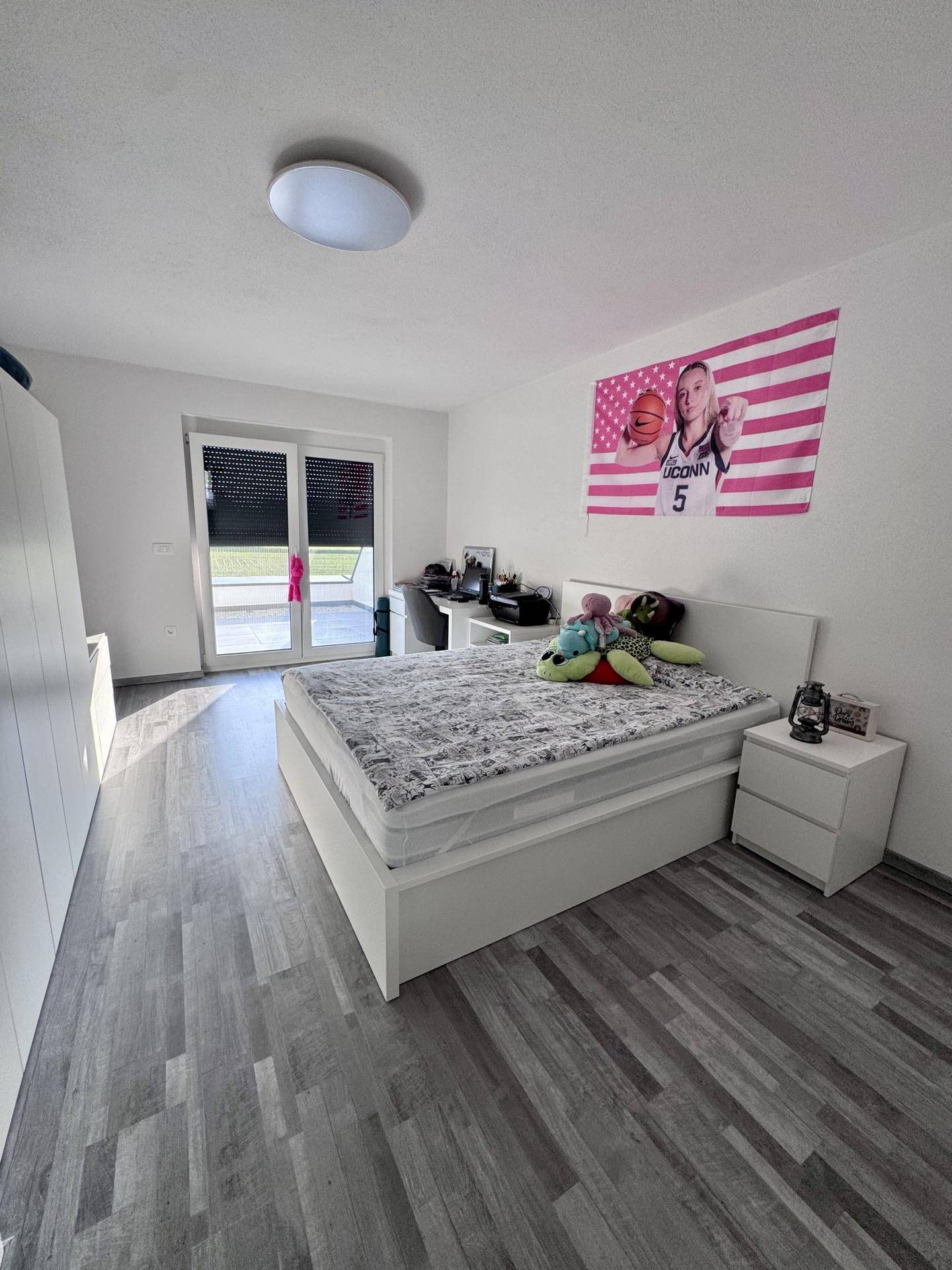 Jugendzimmer mit Bett, Schreibtisch und Laminatboden, hell durch große Fenster.