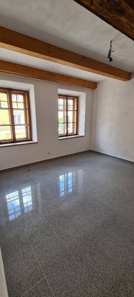 Geräumiger Wohnraum mit sichtbaren Holzbalken, zwei Fenstern und glänzendem Fliesenboden.