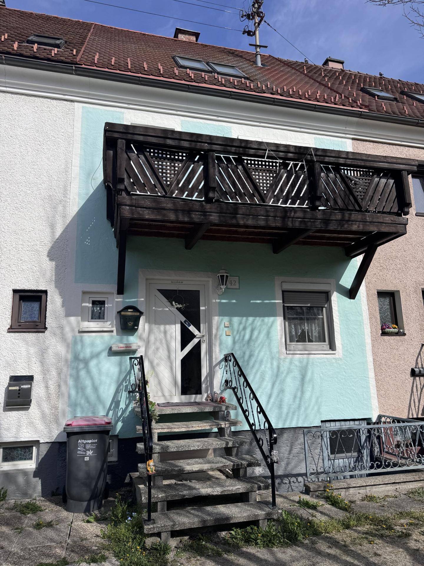 Vorderansicht eines Wohnhauses mit Eingangsbereich, Außentreppe und einem Holzbalkon im Obergeschoss.
