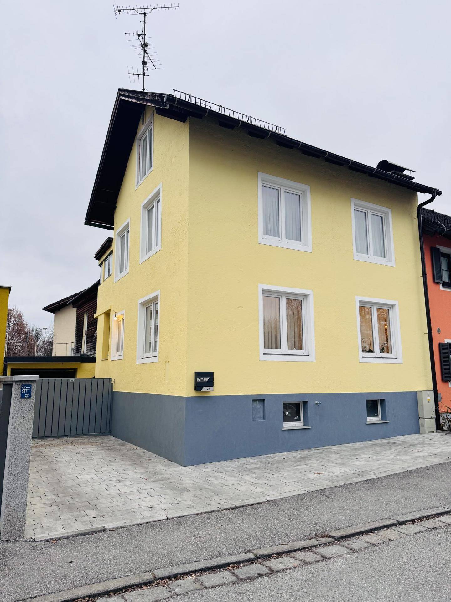 Gelbes Wohnhaus mit grauer Sockelzone, weißen Fenstern und gepflasterter Einfahrt.