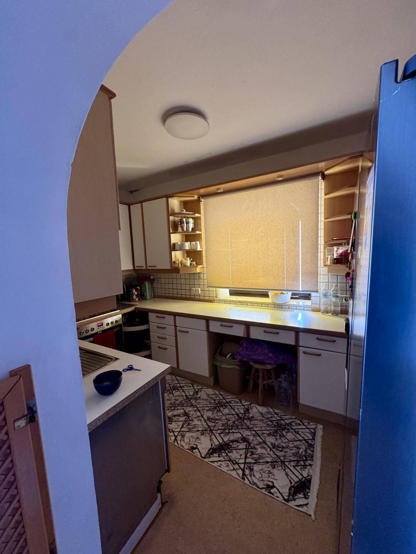 Küche mit Holzschränken, Fenster und Arbeitsbereich, gesehen durch einen Torbogen.