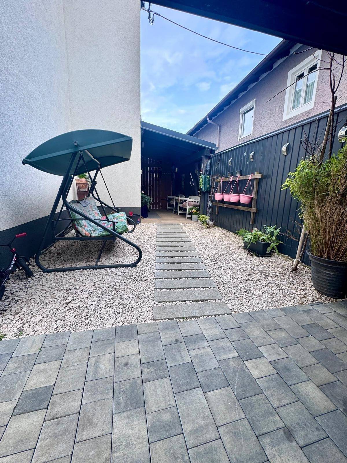 Kleiner Gartenbereich mit Schaukel und gepflastertem Weg, umgeben von einer Holzwand.