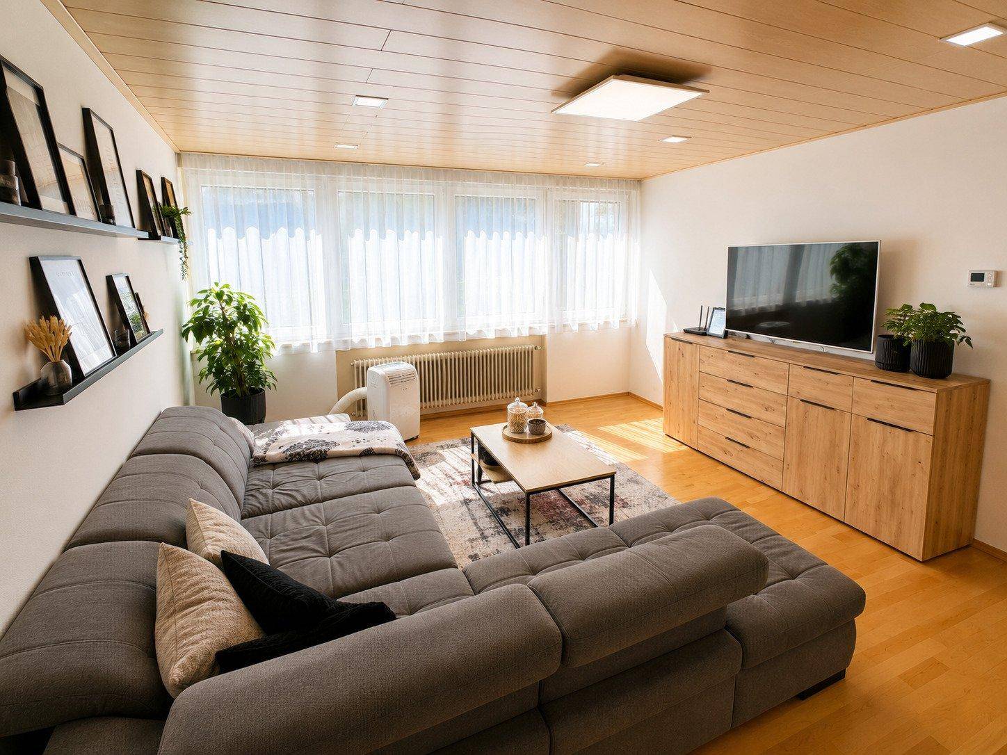 Helles Wohnzimmer mit Holzdecke, großem Ecksofa, Holzboden und Blick nach draußen.
