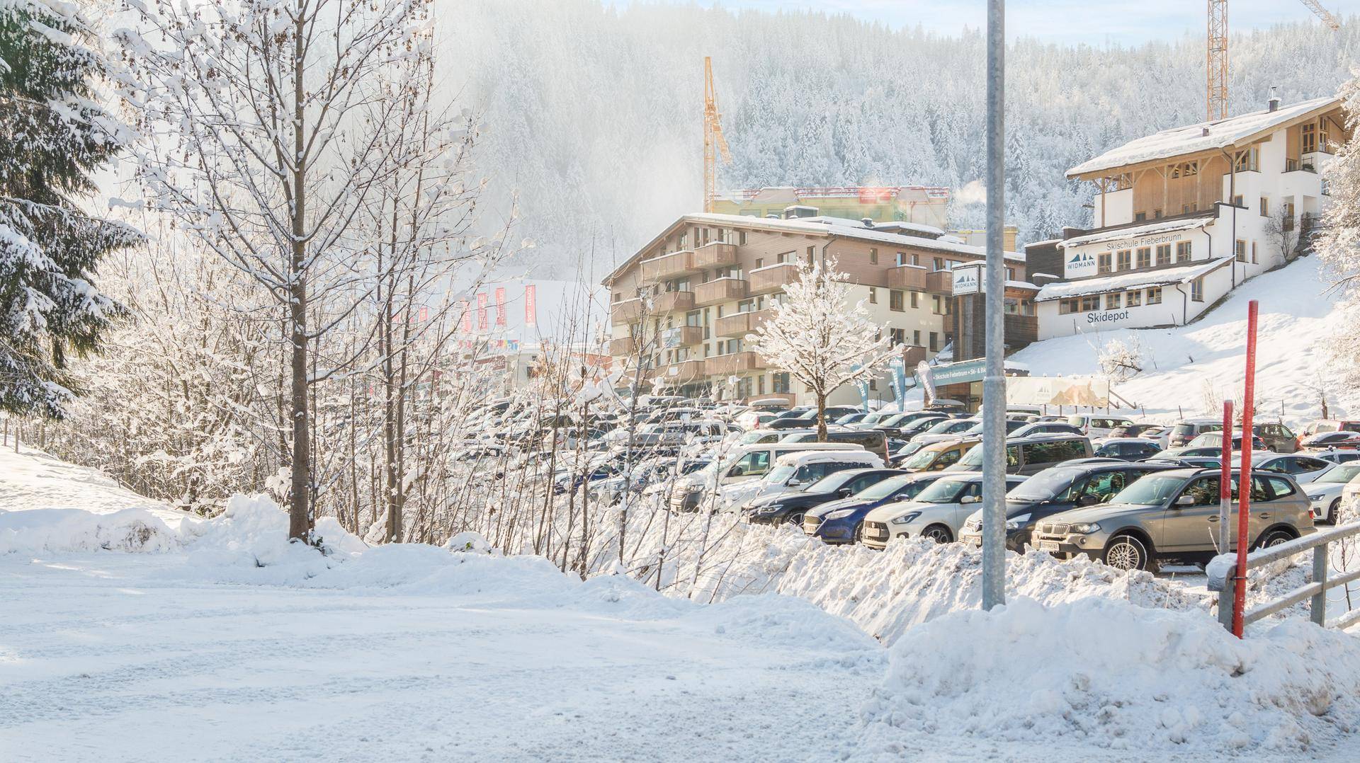 Verschneite Parkplätze vor Gebäuden und bewaldeten Hängen in einer winterlichen Berglandschaft.