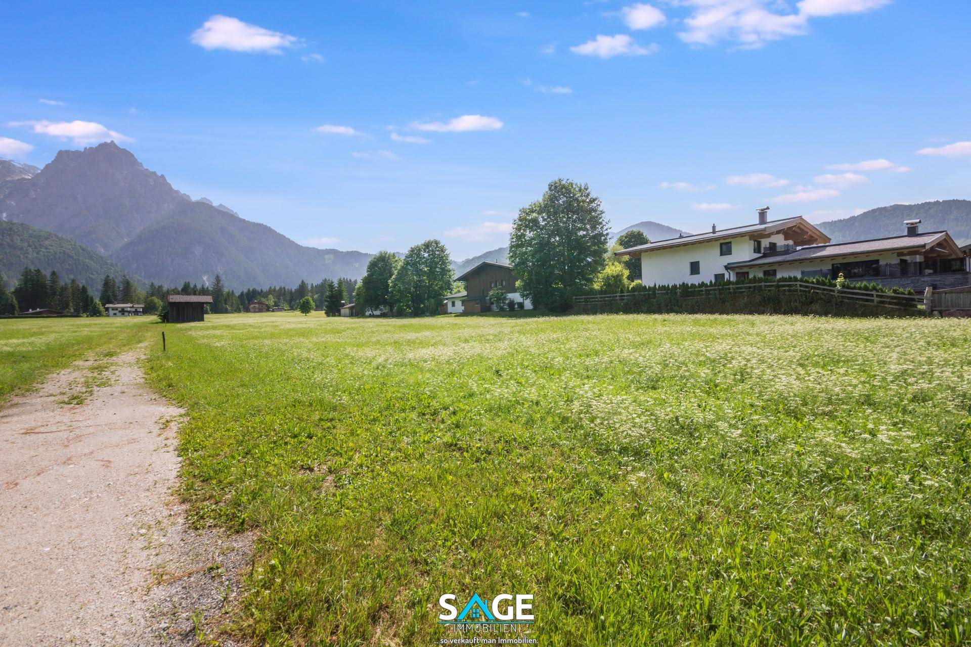 Weitläufige grüne Wiese mit einem Feldweg, umgeben von Häusern und majestätischen Bergen im Hintergrund.
