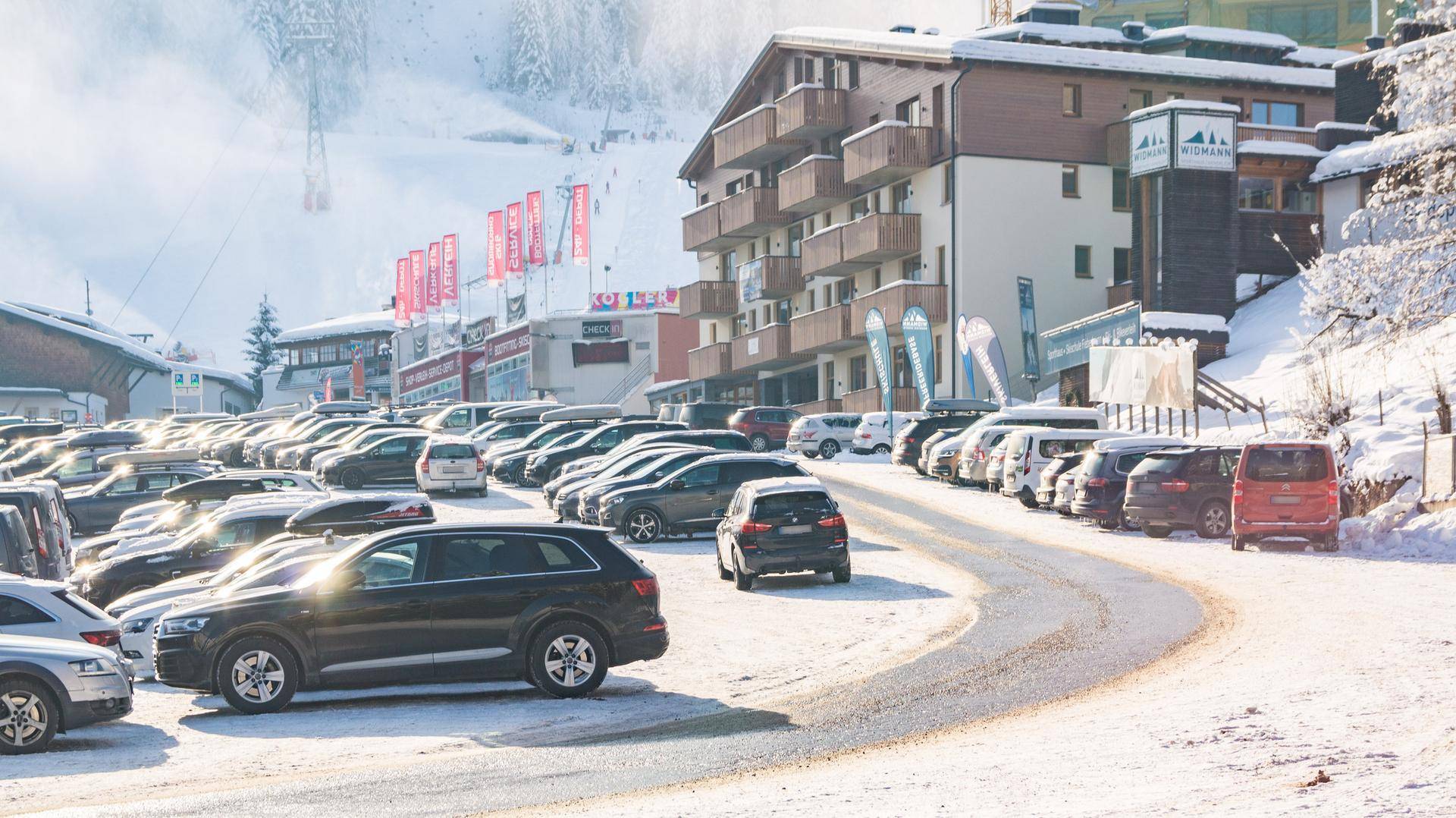 Weitläufiger, verschneiter Parkplatzbereich mit Gebäuden und einer Zufahrtsstraße in den Bergen.