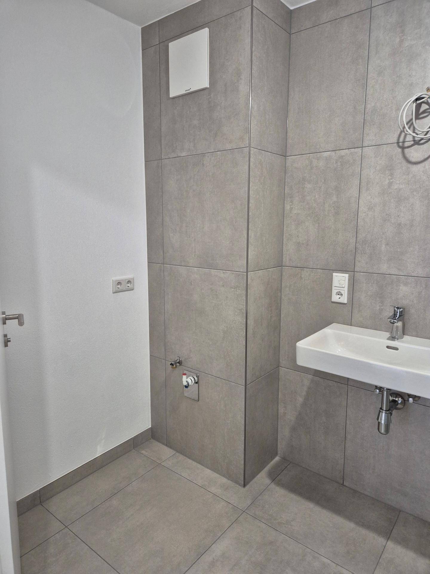 Modernes Gäste-WC mit grauen Fliesen, kleinem Waschbecken und Wand-WC-Anschluss.