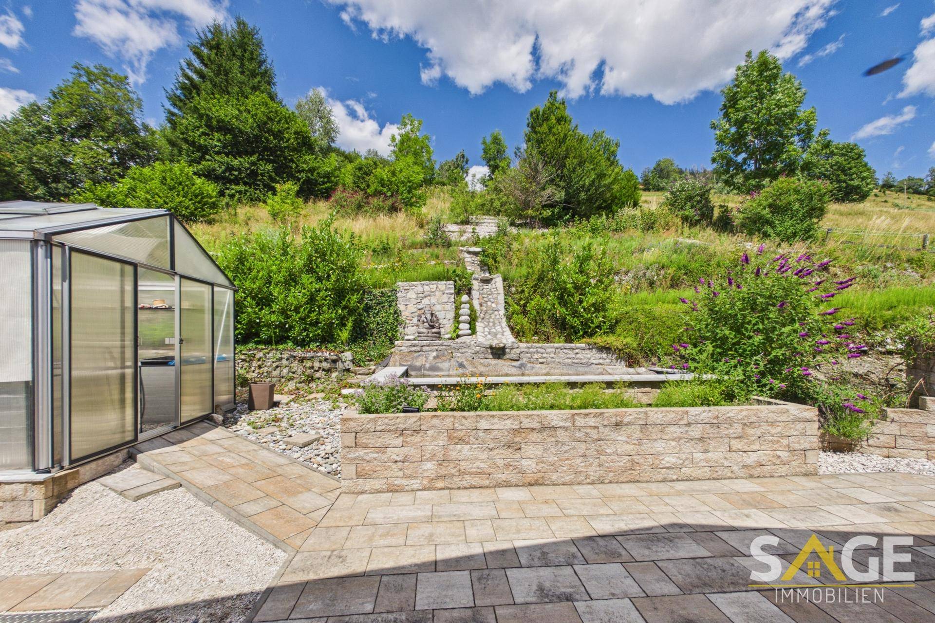 Gepflasterte Terrasse mit Steingarten, Wasserfall und Gewächshaus, umgeben von üppiger Natur.