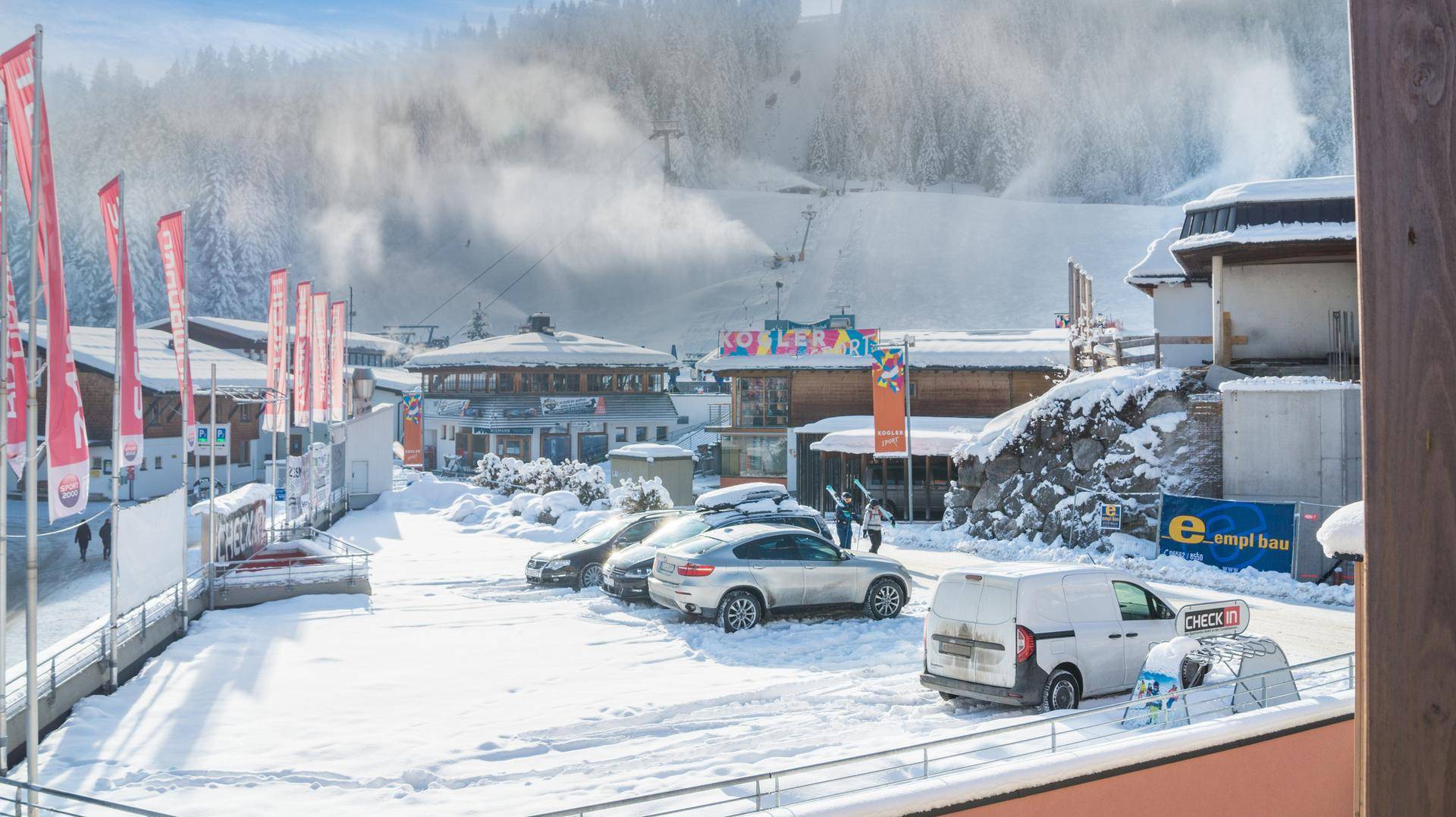 Winterliche Landschaft mit verschneiten Gebäuden, Parkplätzen und einer Skipiste im Hintergrund.