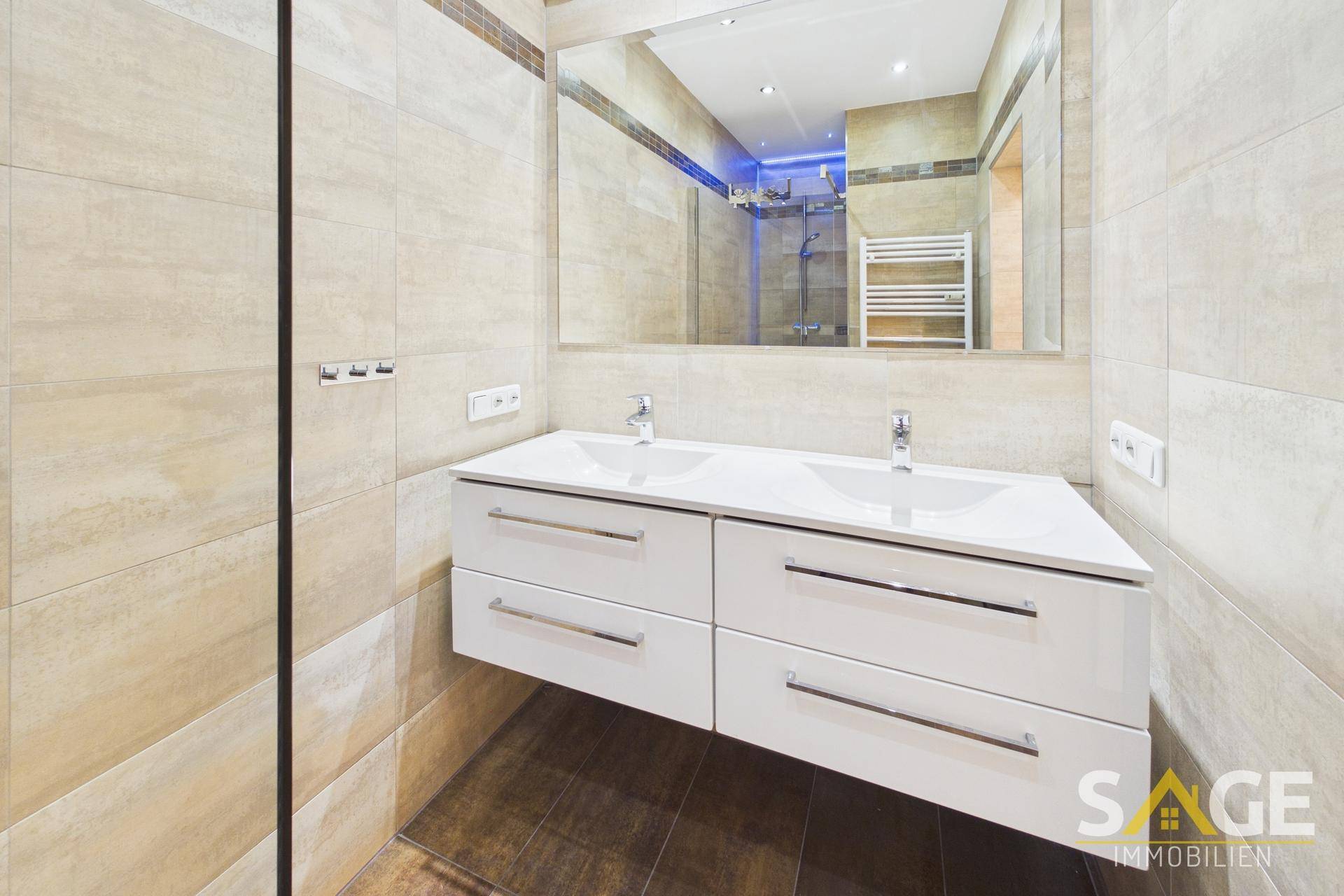 Modernes Badezimmer mit Doppelwaschbecken, großem Spiegel und beigefarbenen Fliesen an Wänden und Boden.