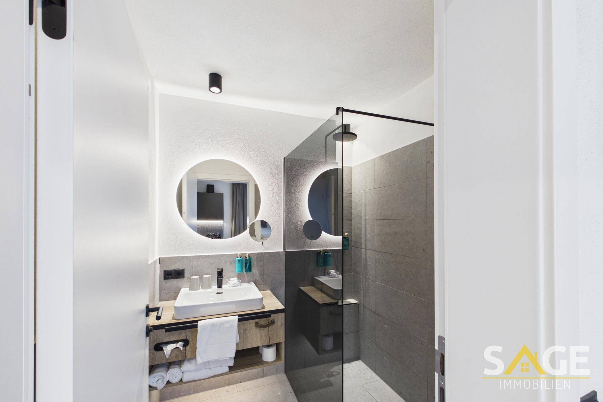 Modernes Badezimmer mit begehbarer Dusche, beleuchtetem Spiegel und grauen Fliesen, sehr stilvoll gestaltet.