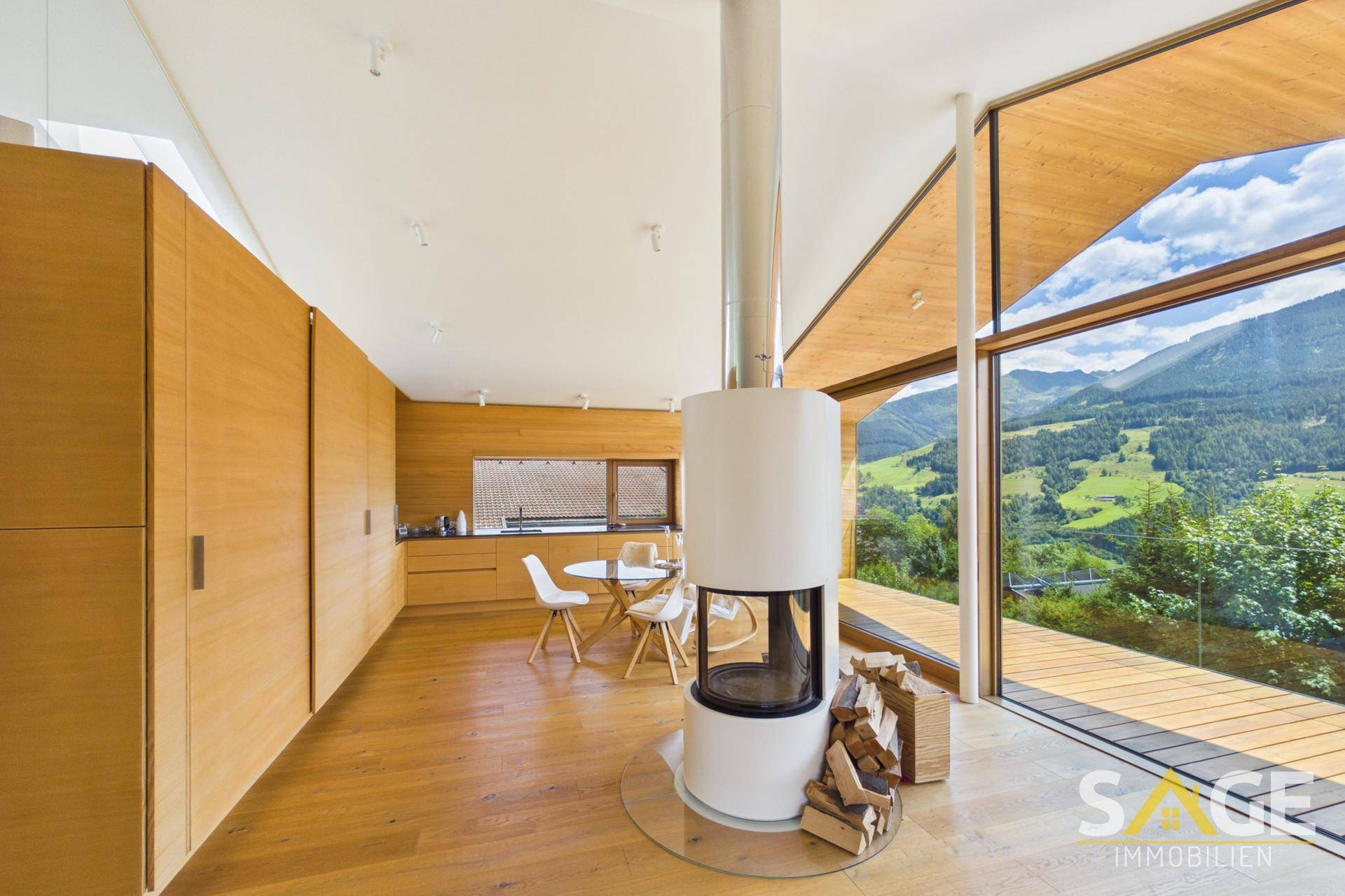 Offener Wohnbereich mit integrierter Küche, Holzelementen und einem zentralen Kamin mit Bergblick.
