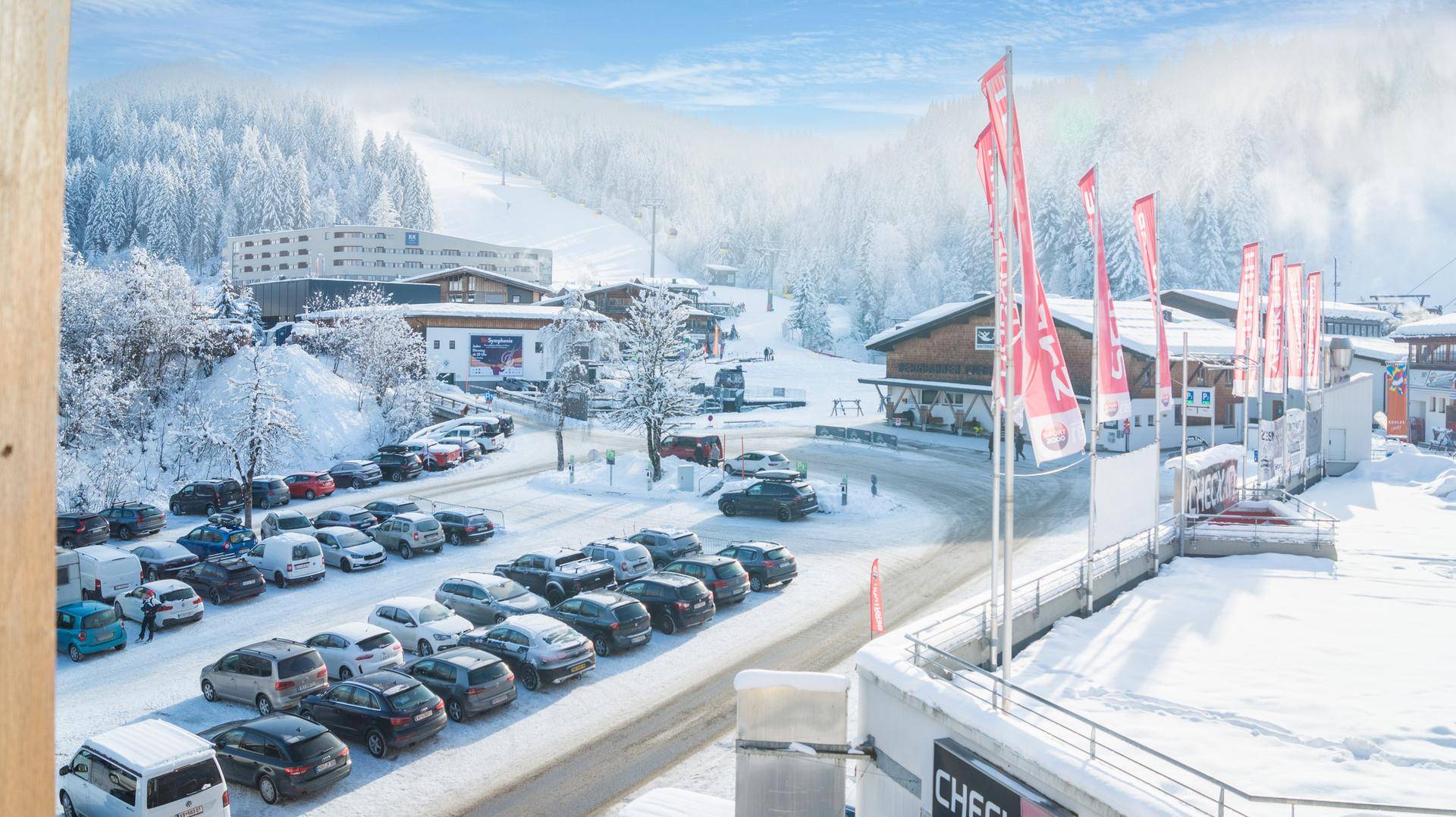 Panoramablick auf das verschneite Skigebiet mit Gebäuden, Parkplätzen und Skipisten unter blauem Himmel.