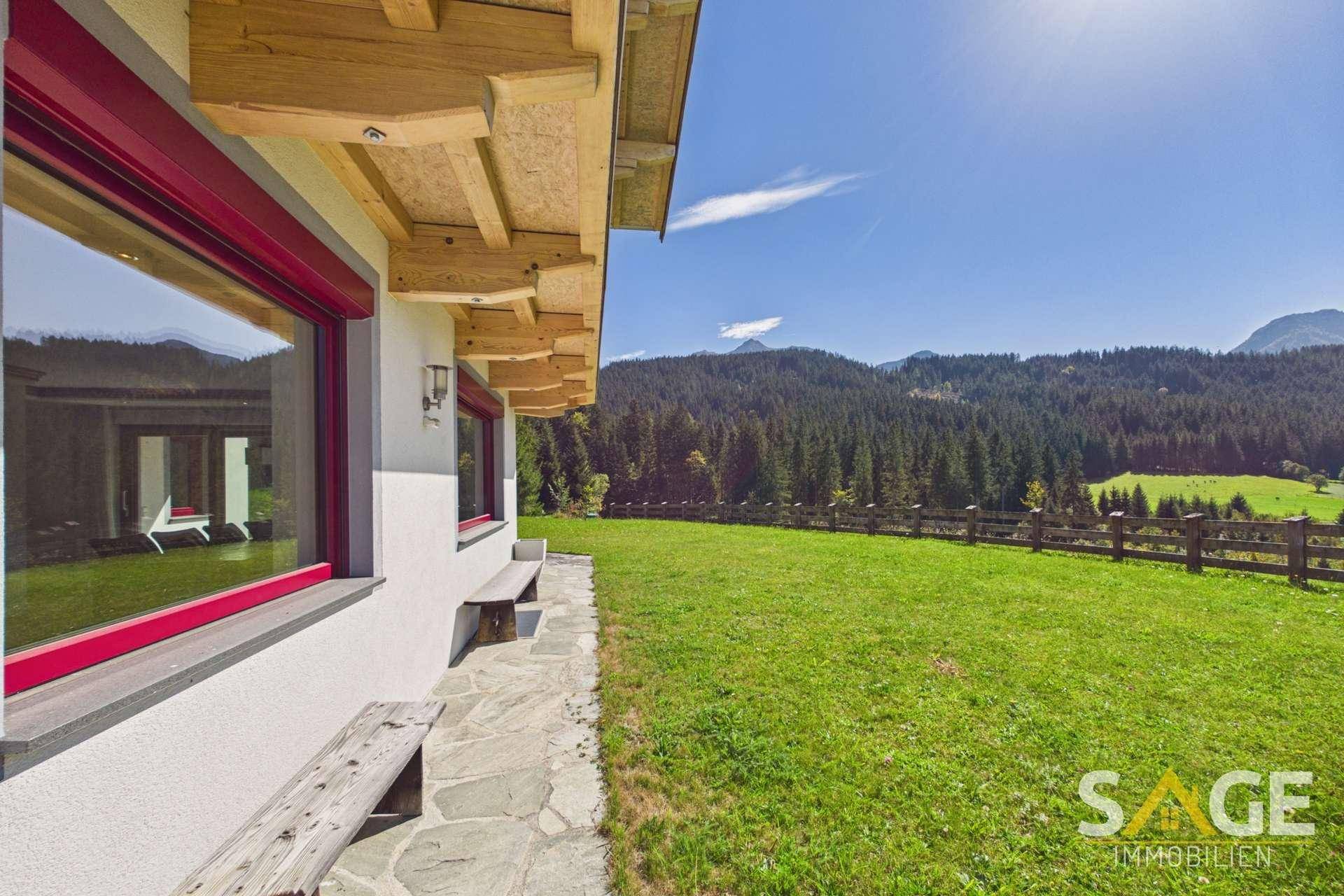 Gebäudefassade mit roten Fensterrahmen, Steinweg und Holzbänken, umgeben von grüner Natur.