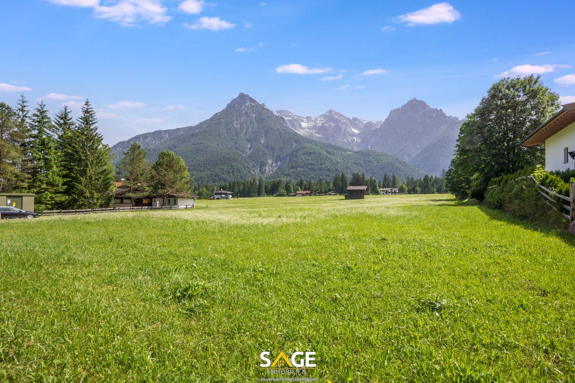 Panoramablick über eine weite grüne Wiese mit Häusern am Rand und hohen Bergen unter blauem Himmel.