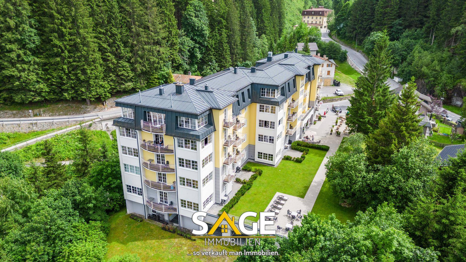 Luftaufnahme eines modernen Apartmentgebäudes mit gelber Fassade, umgeben von dichtem Wald und Bergen.