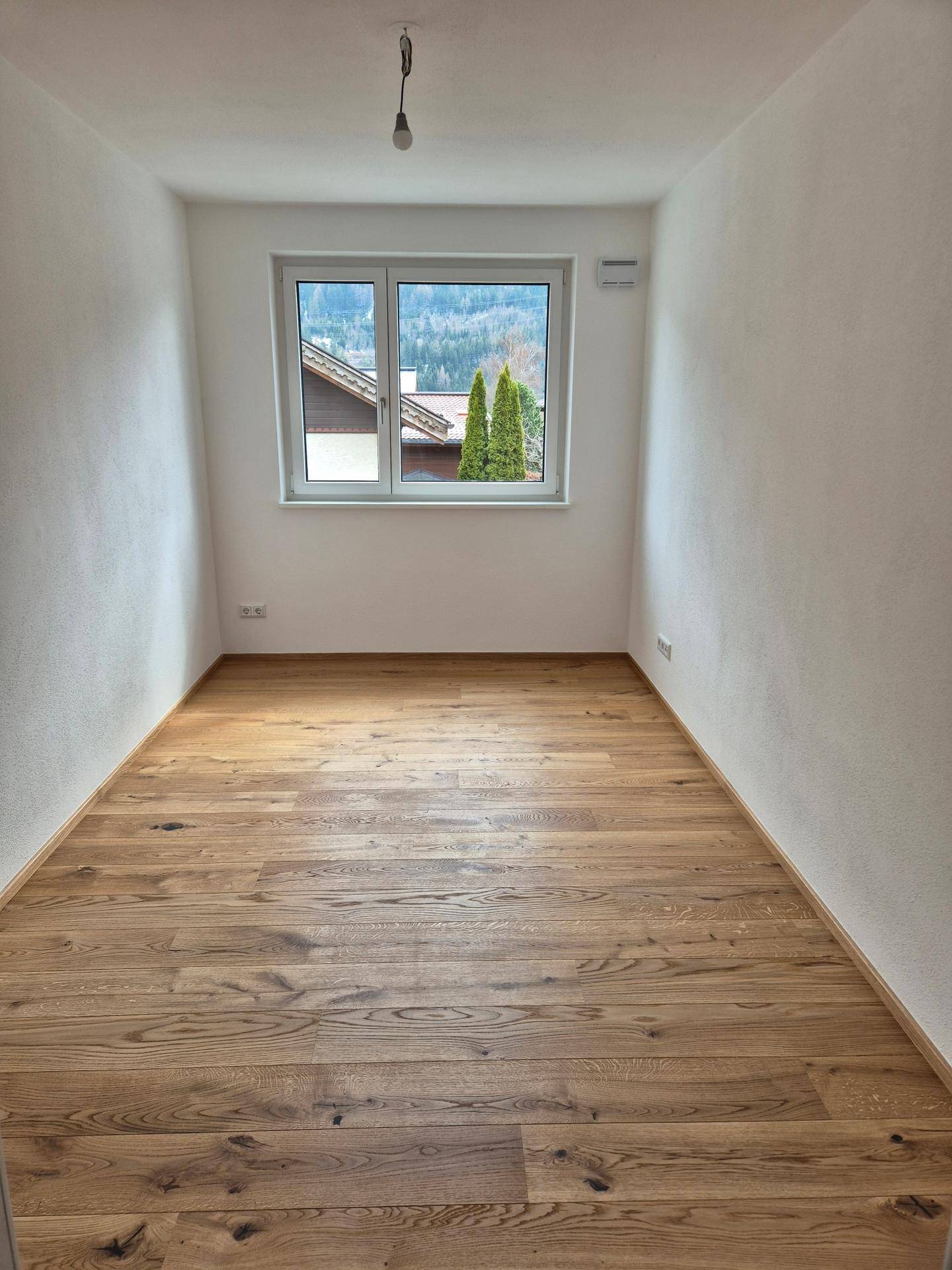 Kleines, helles Zimmer mit Holzboden und Fensterblick auf Bäume und die bergige Landschaft.