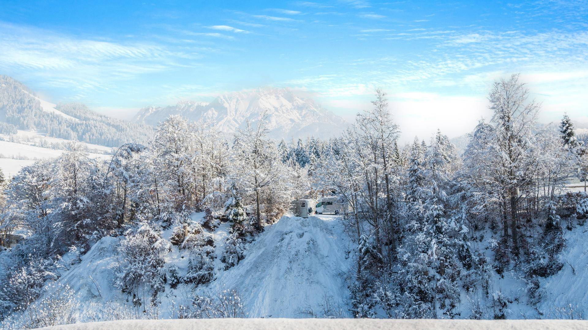Weitläufige, verschneite Berglandschaft mit dichten Wäldern und einem Wohnmobil unter blauem Himmel.