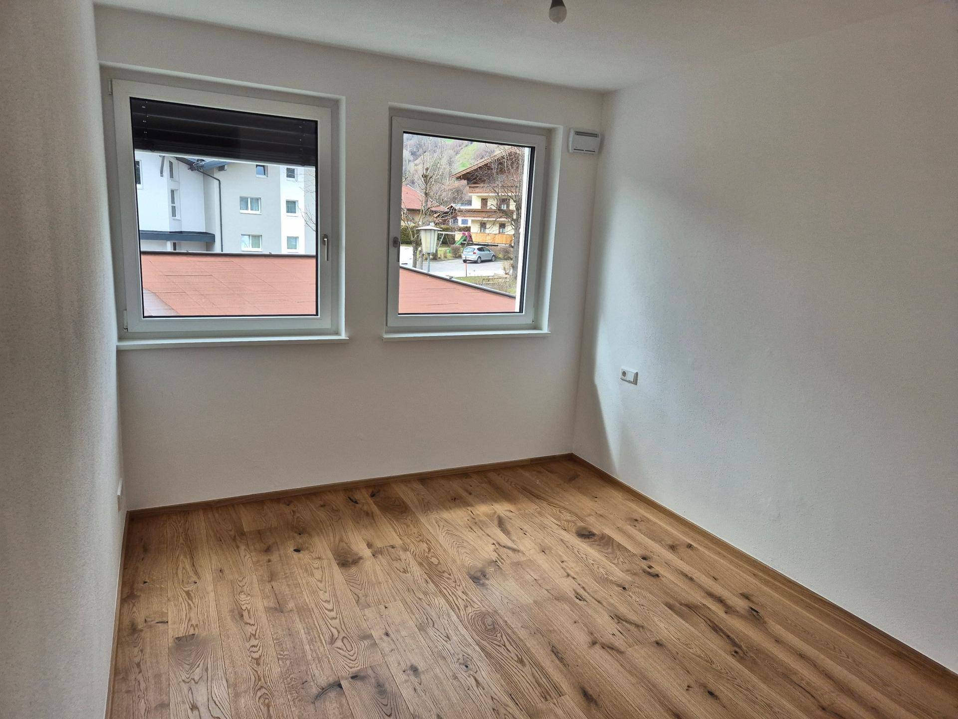 Helles Zimmer mit Holzboden und zwei Fenstern, die auf die Umgebung blicken.
