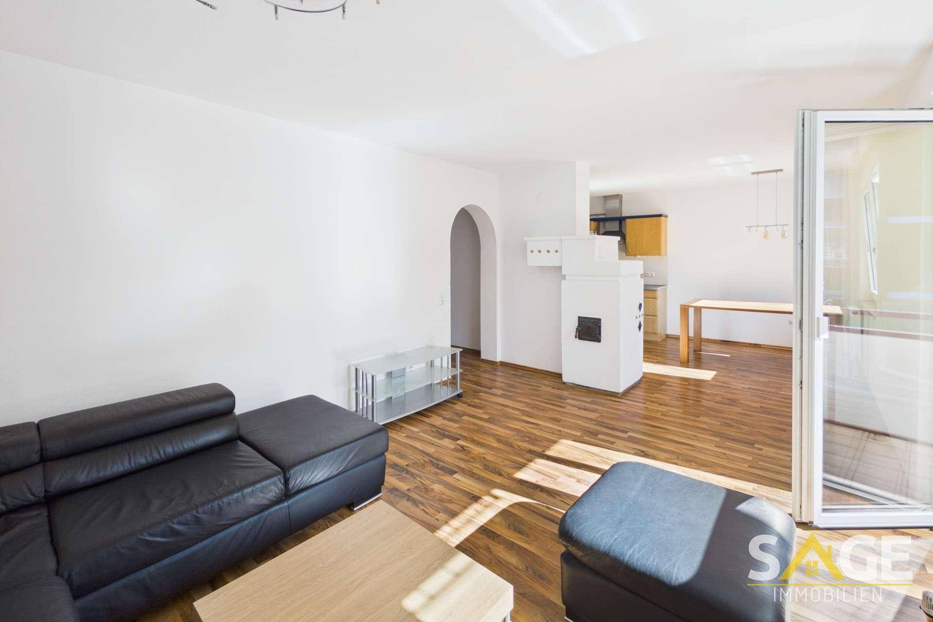 Heller Wohnbereich mit Holzboden, schwarzem Sofa und traditionellem Kachelofen.