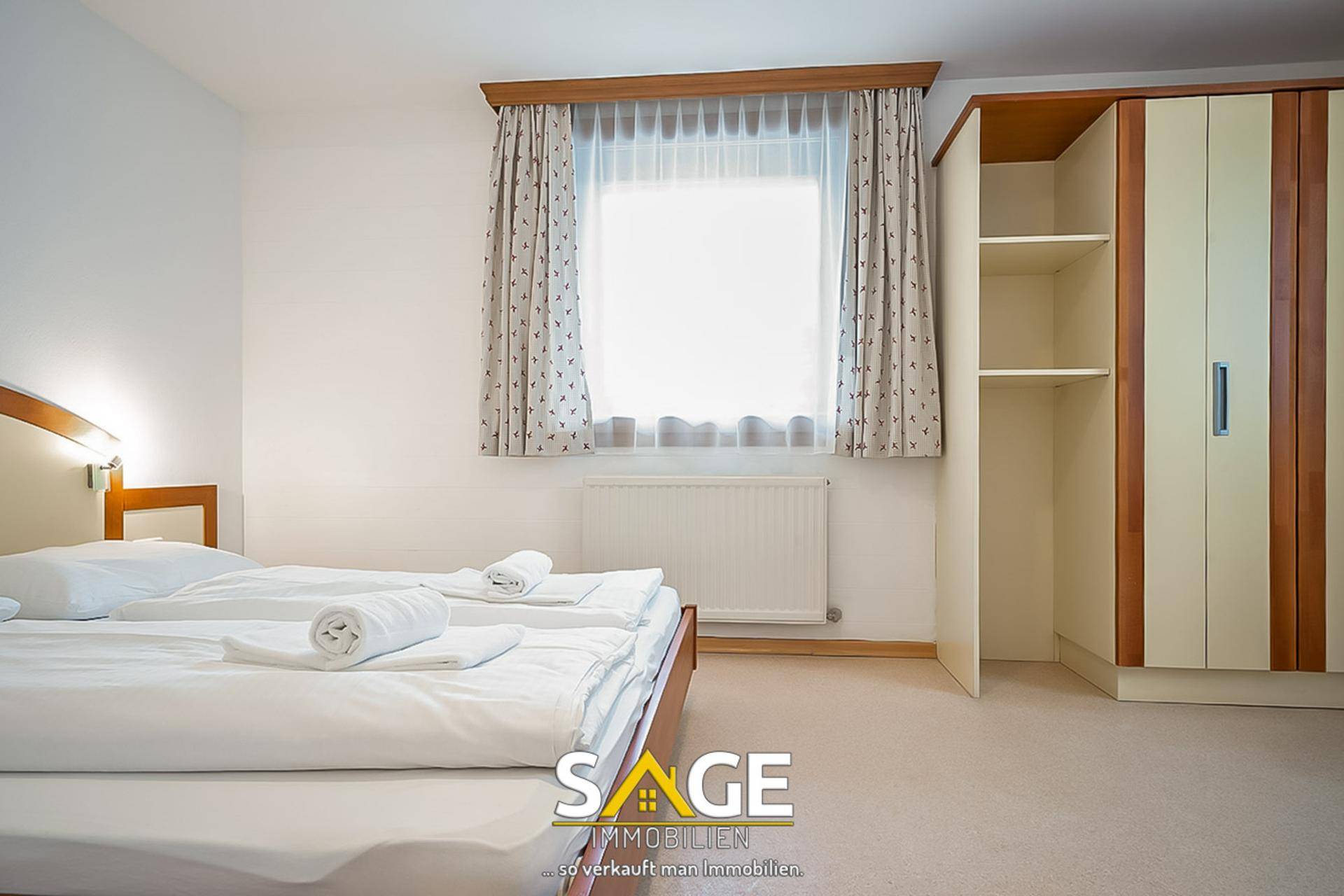 Helles Schlafzimmer mit Doppelbett, Fenster und einem Kleiderschrank in Holzoptik.
