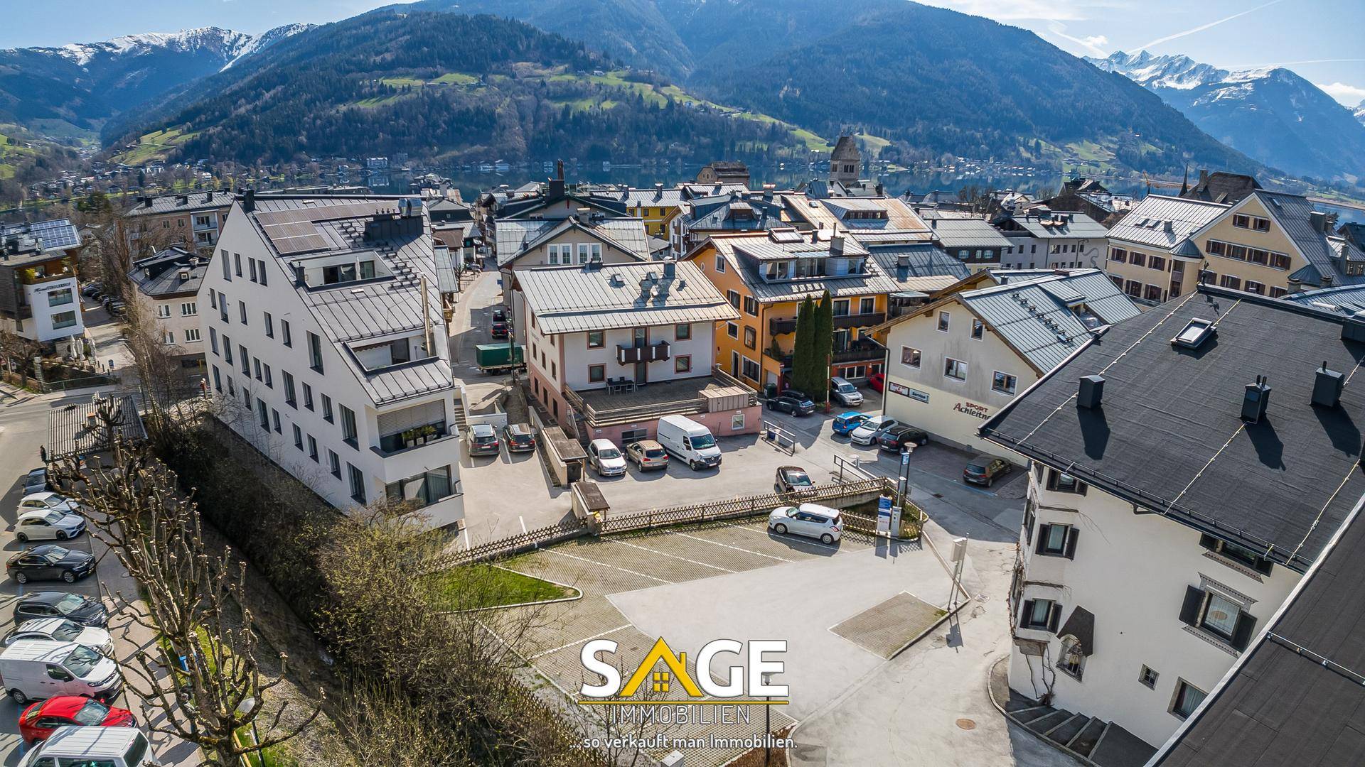 Luftaufnahme einer städtischen Siedlung mit verschiedenen Gebäuden und Parkplätzen, umgeben von Bergen.