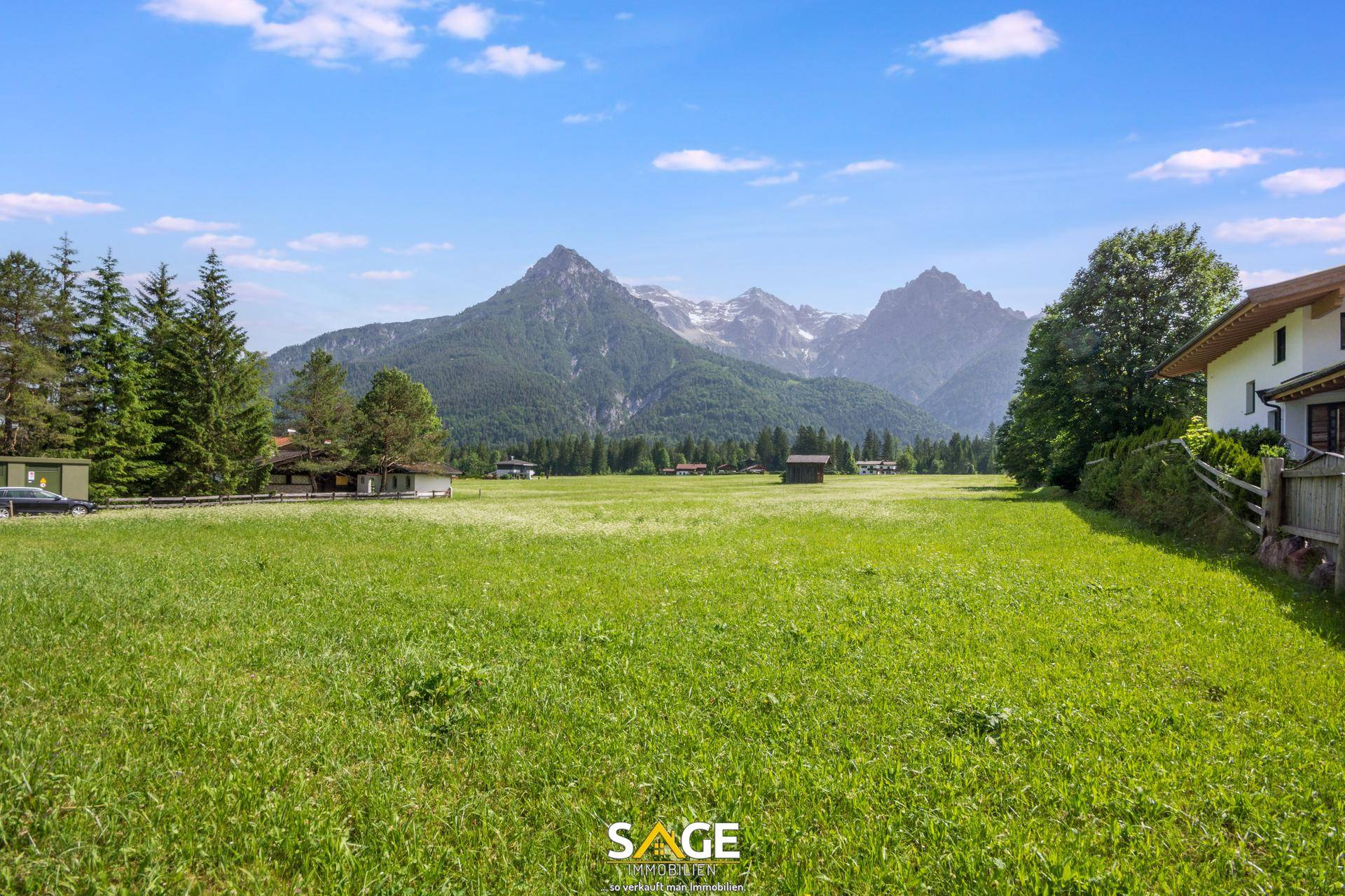 Ausgedehnte grüne Wiesenfläche mit vereinzelten Häusern und einer beeindruckenden Bergkette im Hintergrund.