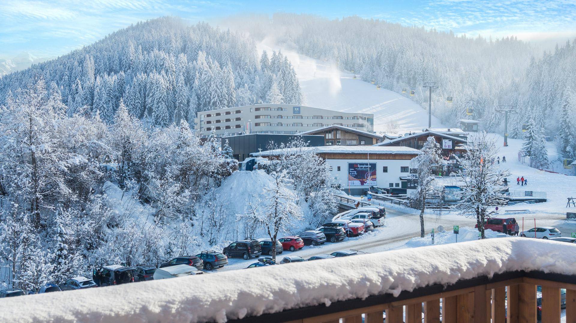 Blick vom Balkon auf das verschneite Skigebiet mit Gebäuden, Parkplätzen und Skipisten.
