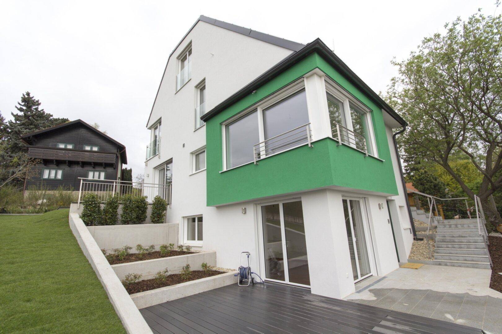 Moderne Villa mit weißer Fassade, grünem Anbau, Holzterrasse und Gartenbereich.