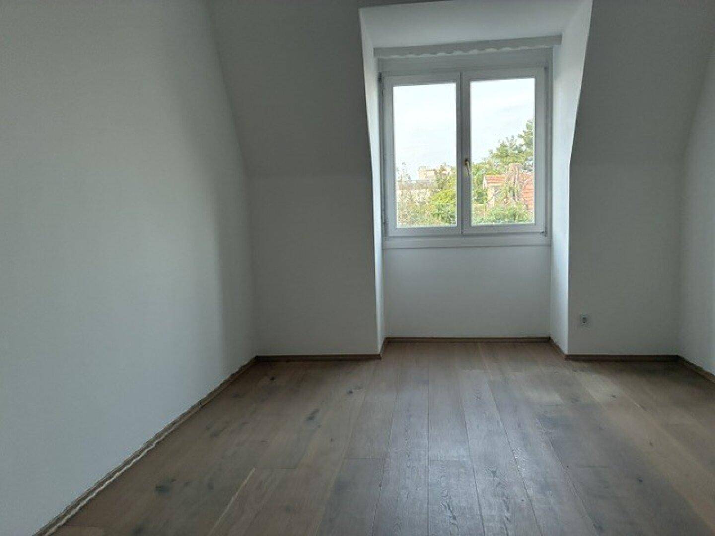Leeres Schlafzimmer mit dunklem Holzboden, Dachschräge und großem Fenster für viel Licht.