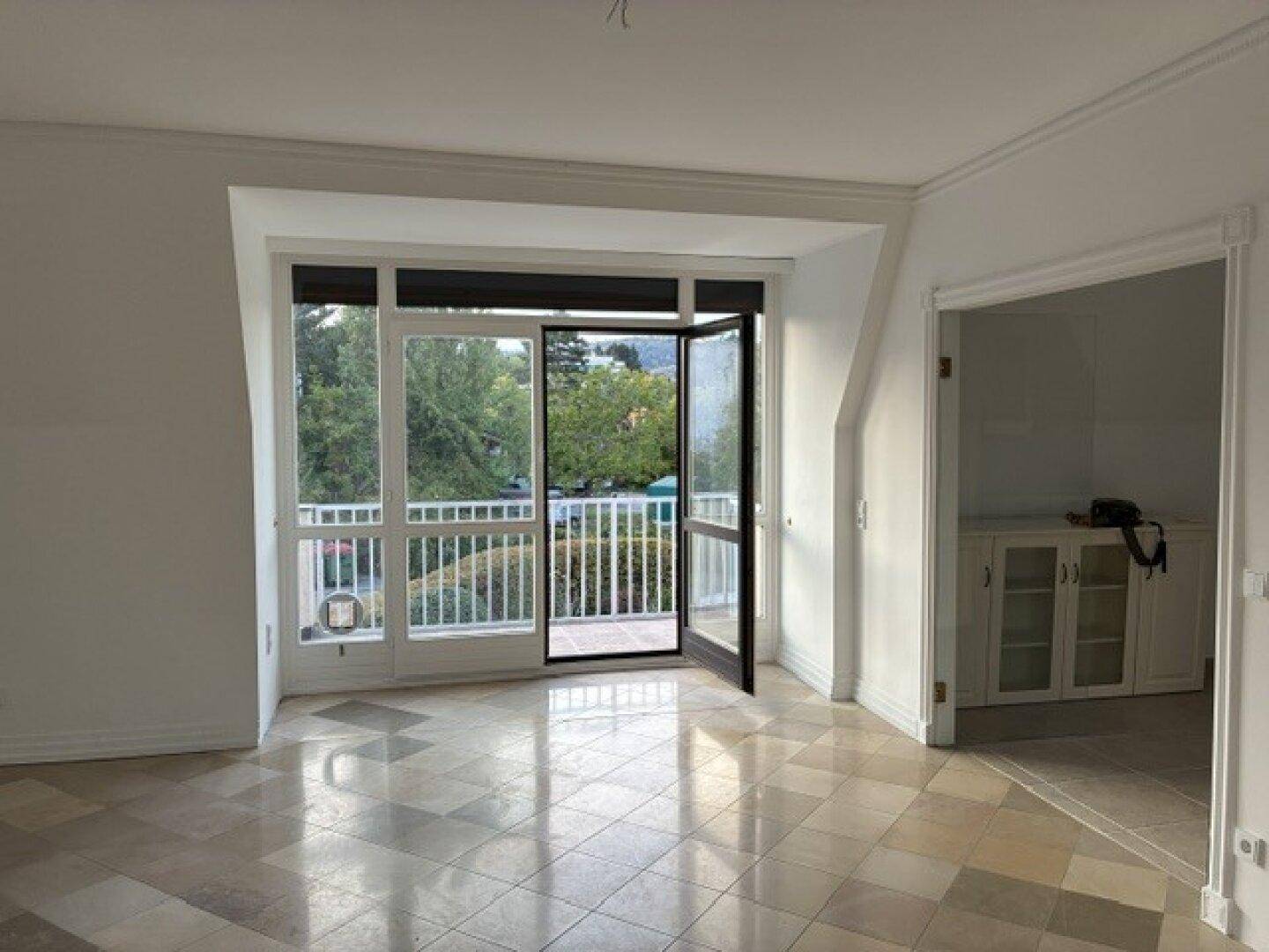 Heller Wohnraum mit Fliesenboden und breiter Fensterfront mit Zugang zum Balkon.