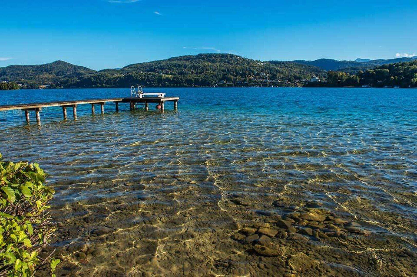 Idyllischer See mit Holzsteg und klarem Wasser, umgeben von grünen Bergen unter blauem Himmel.
