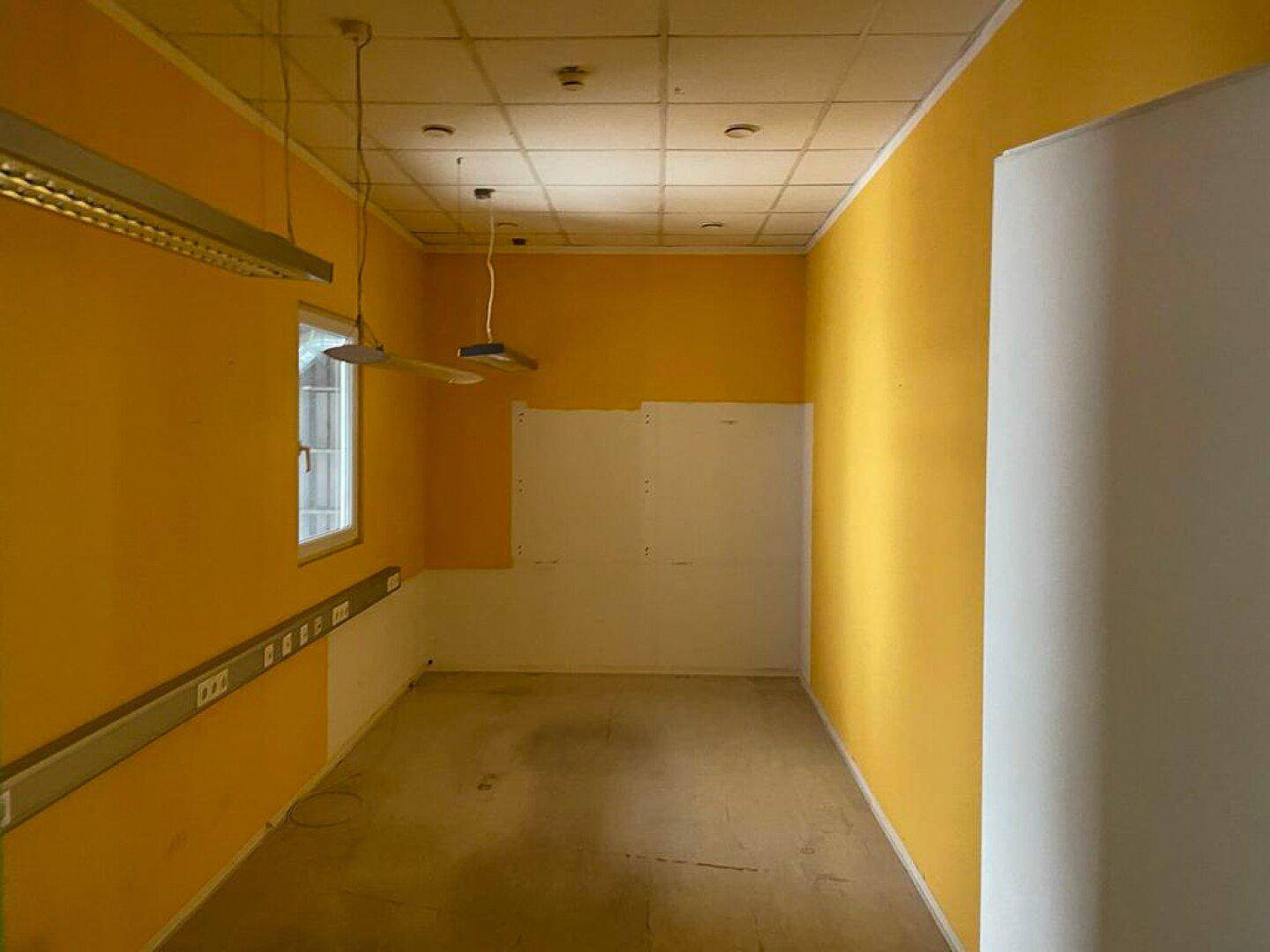 Kleiner Büroraum mit leuchtend gelben Wänden, abgehängter Decke und einem Fenster.