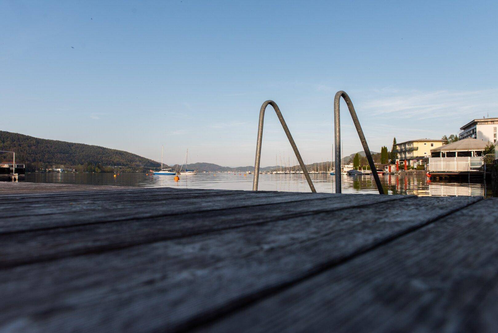 Holzsteg mit Badeleiter am Seeufer, umgeben von Bergen und Gebäuden, mit Blick auf Segelboote.
