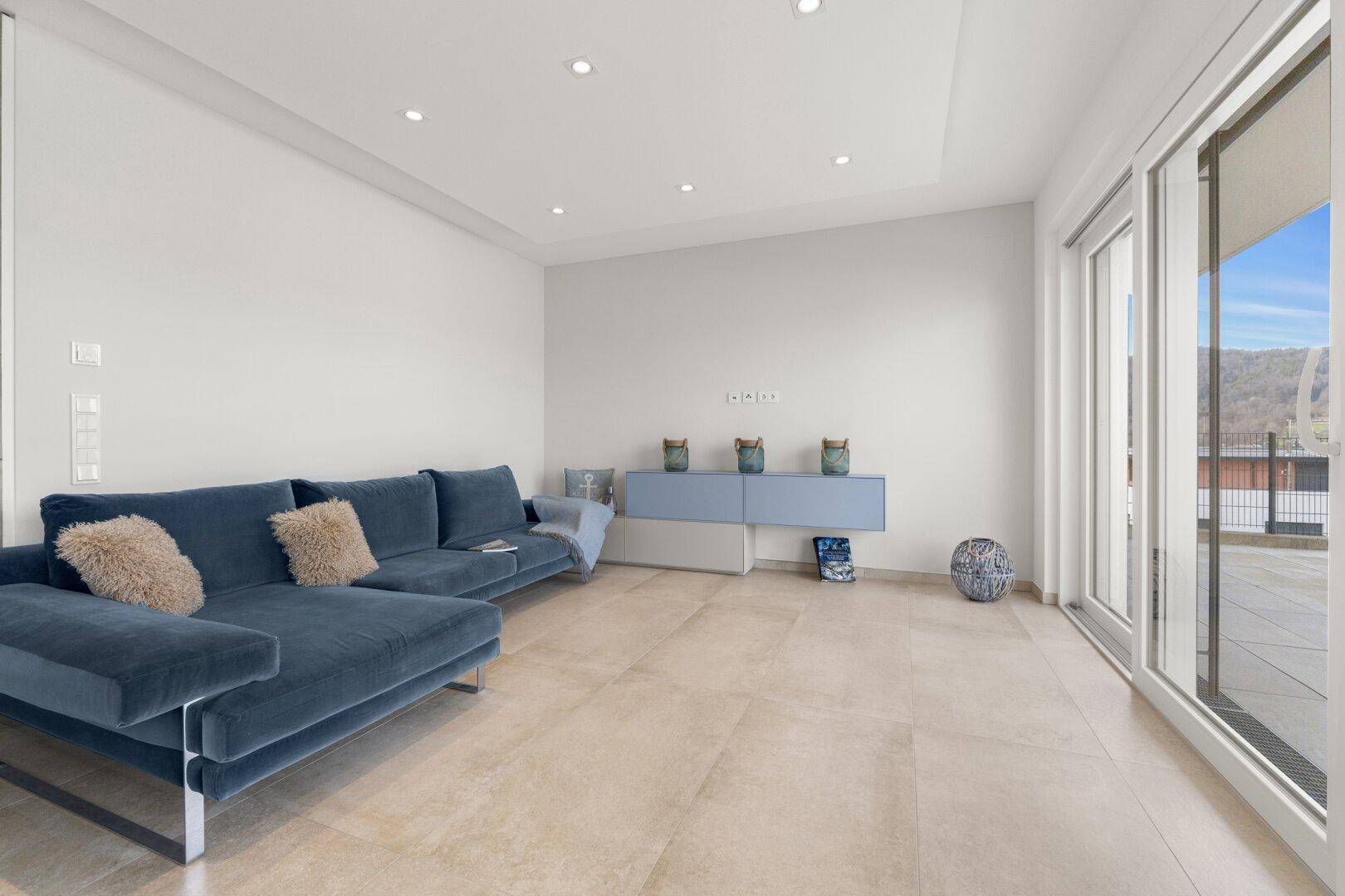 Helles Wohnzimmer mit bequemer blauer Couch, minimalistischer Einrichtung und Zugang zur Terrasse.