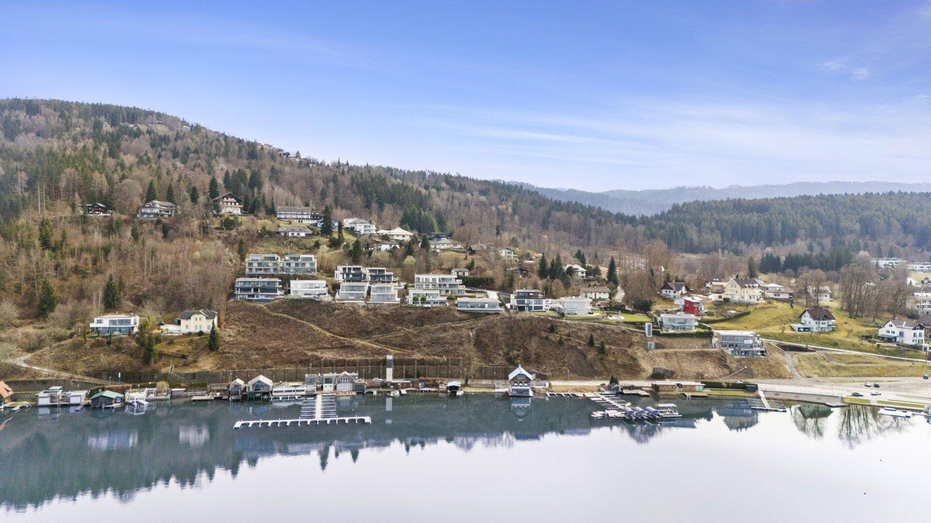 Panoramablick auf den See mit umliegenden Häusern, bewaldeten Hügeln und klarem Himmel.