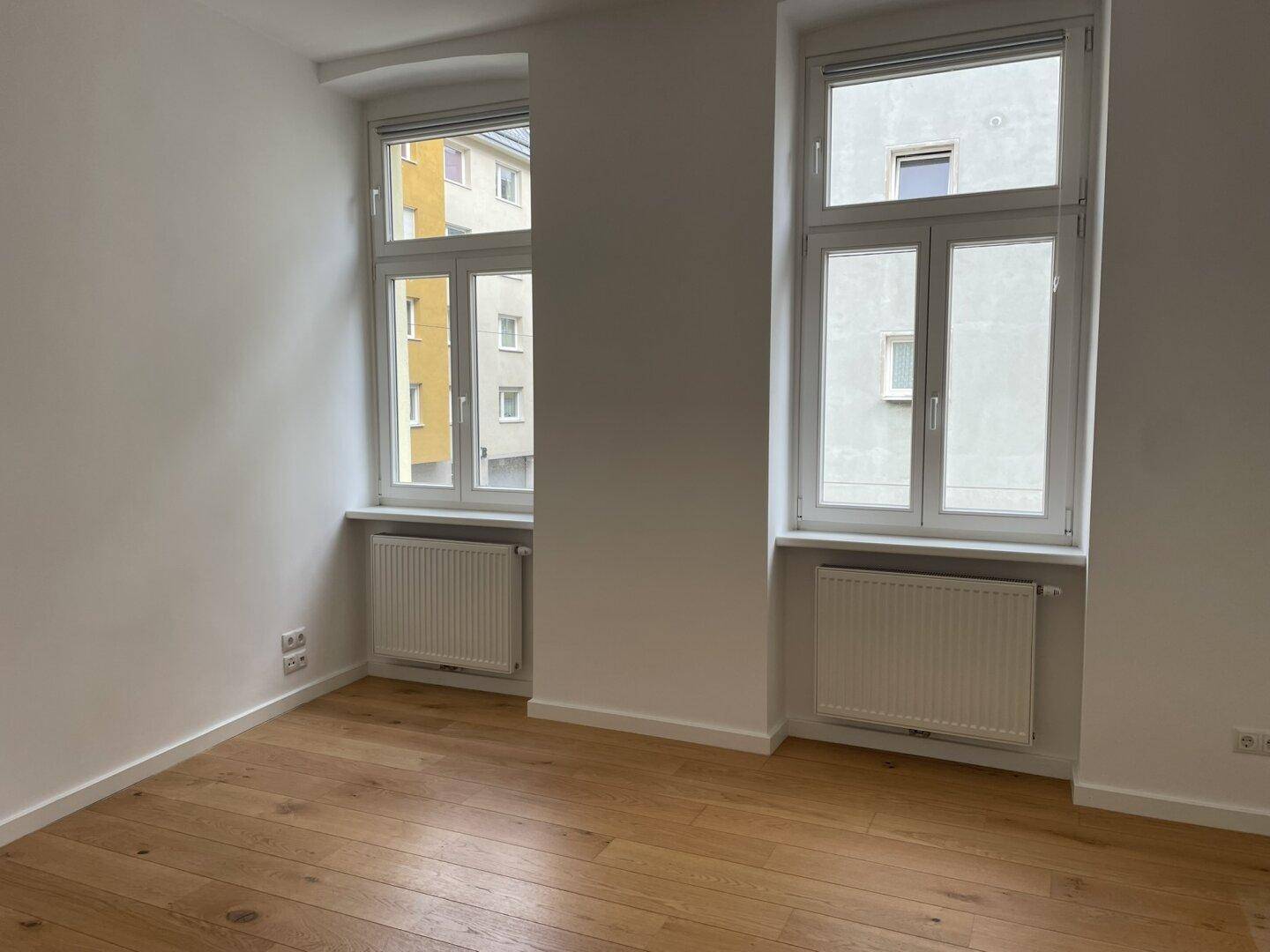 Helles Schlafzimmer mit Parkettboden und zwei großen Fenstern.
