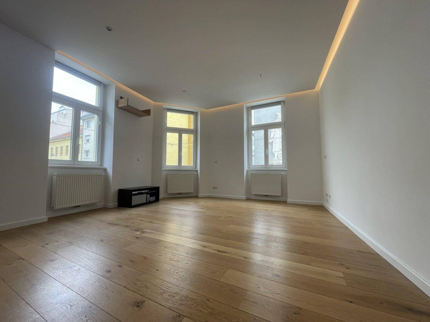 Geräumiges Wohnzimmer mit Parkettboden, großen Fenstern und indirekter Deckenbeleuchtung.