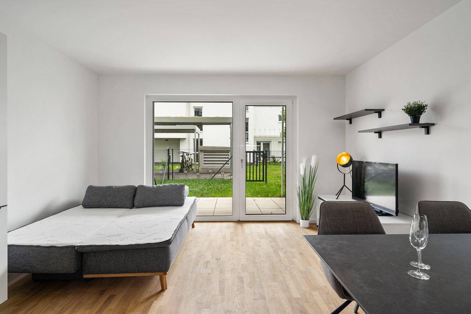 Heller Wohnbereich mit Schlafsofa und Essplatz, bietet direkten Zugang zu einer kleinen Grünfläche.
