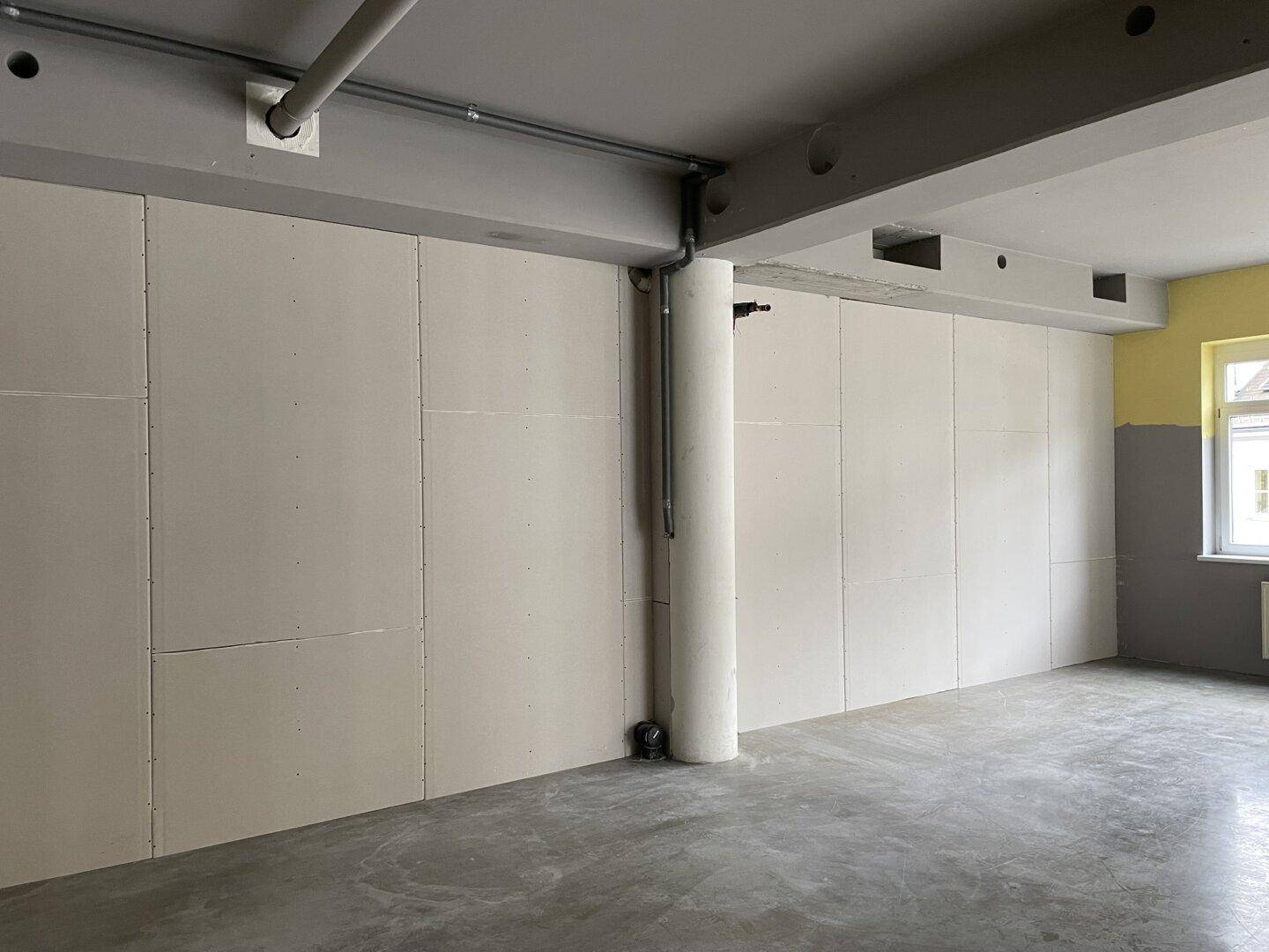 Langer Innenraum mit Trockenbauwänden, einem Fenster und sichtbaren Deckenbalken und Rohren.